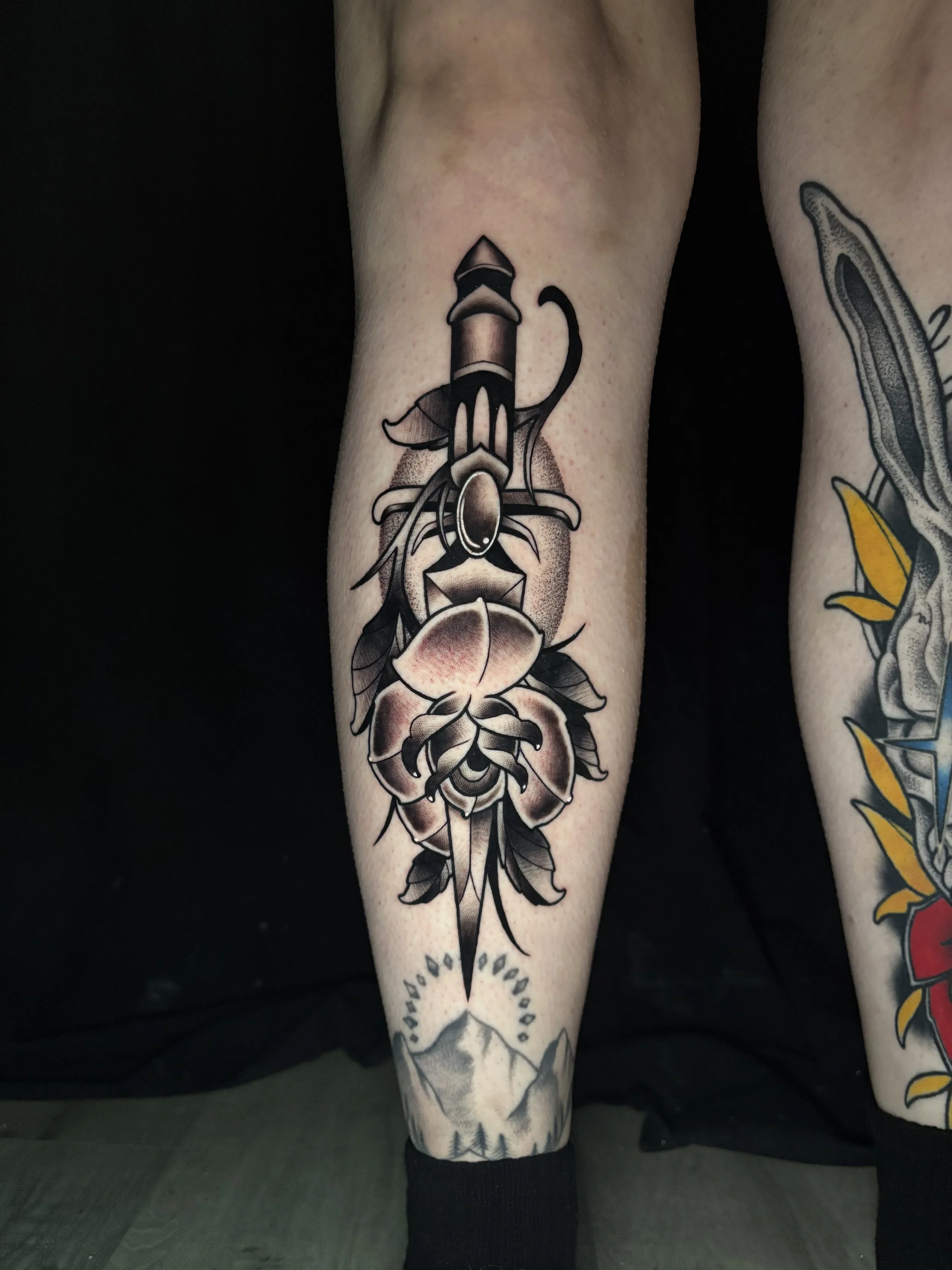 Tattoo of a dagger with a rose at the bottom and decorative elements on the person's leg.