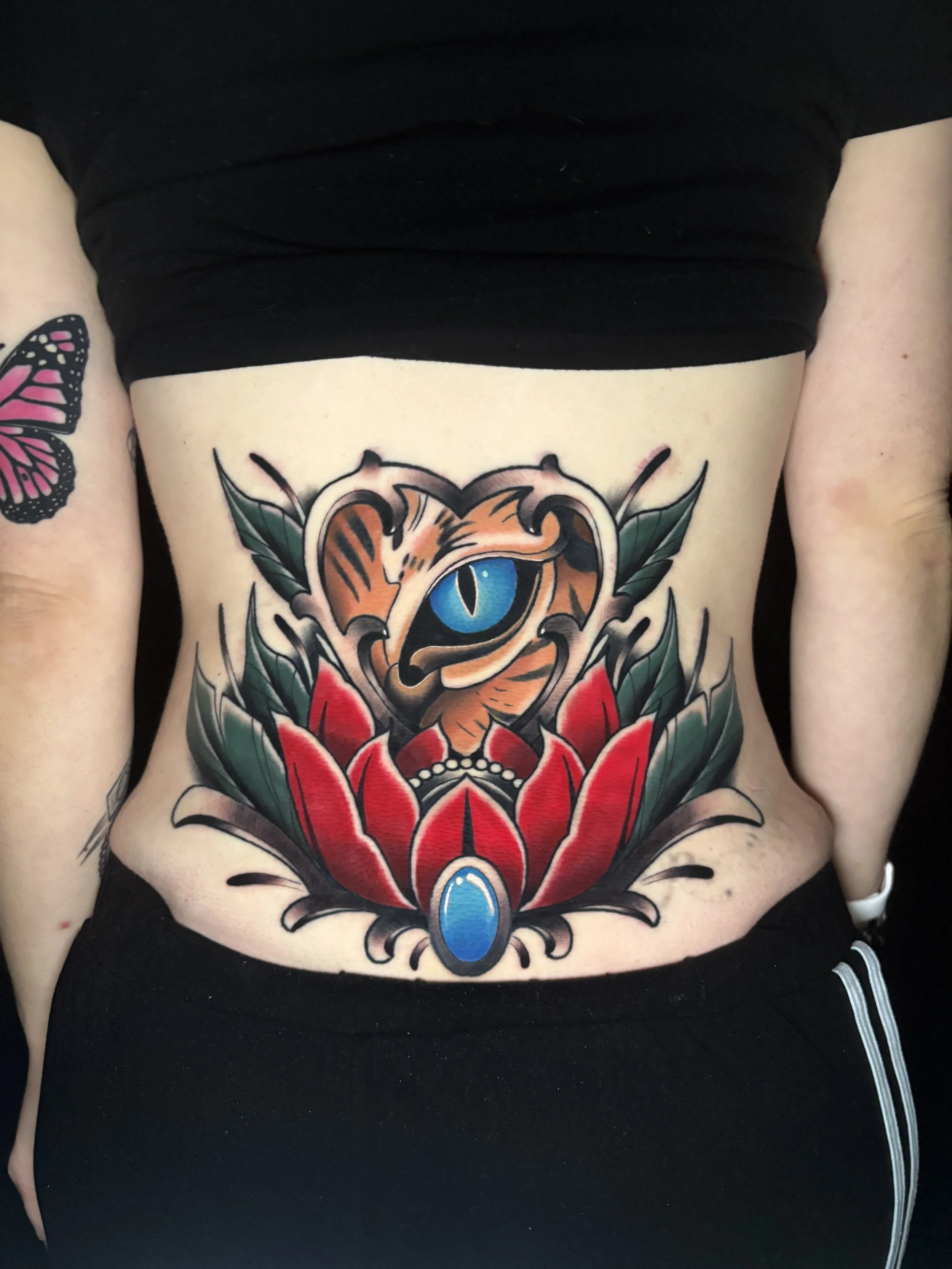 Colorful tattoo of a tiger's face with blue eyes, surrounded by green leaves and red flowers, located on the person's lower back.