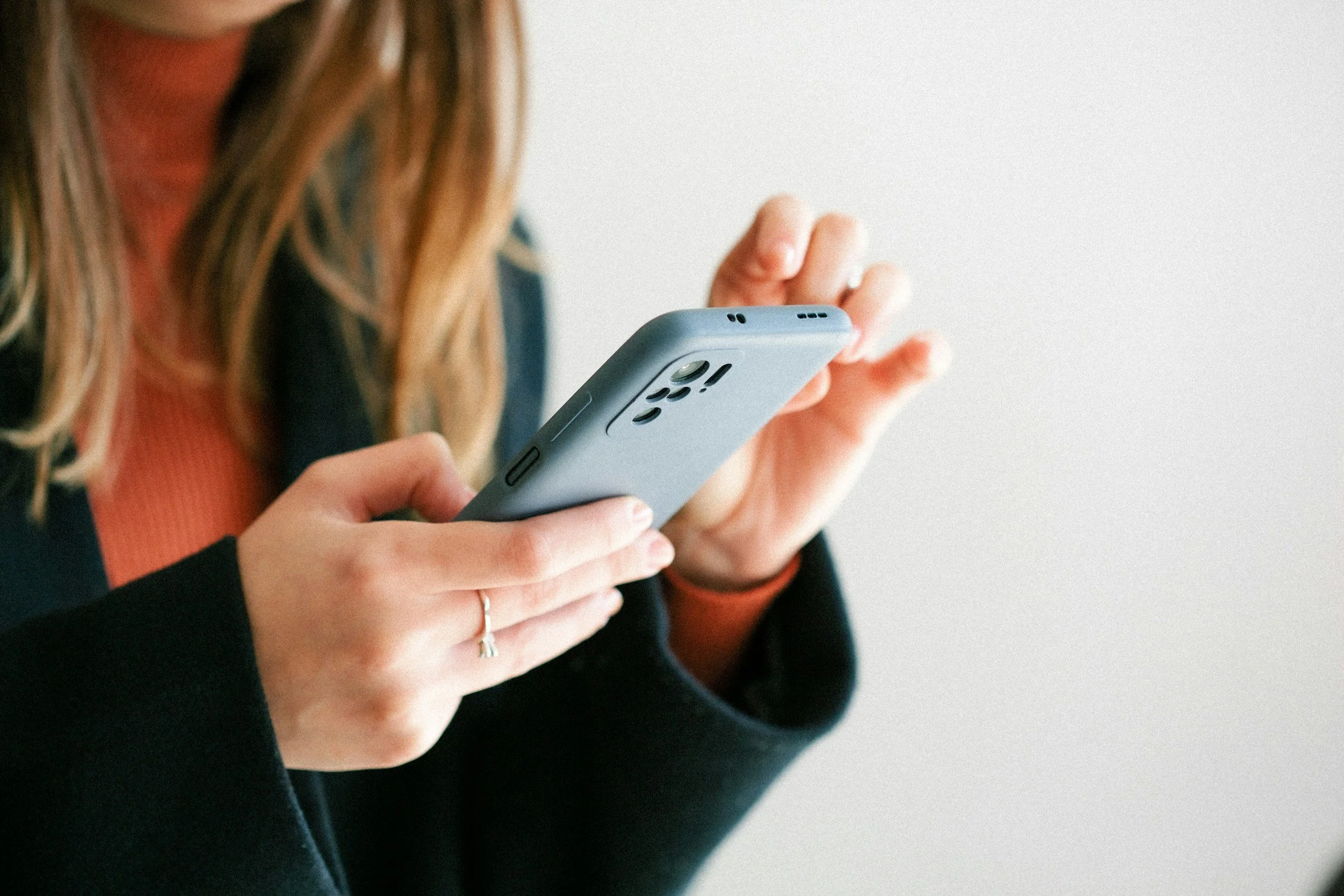 A person holding a smartphone with both hands, with the right hand typing on it, against a plain light background.