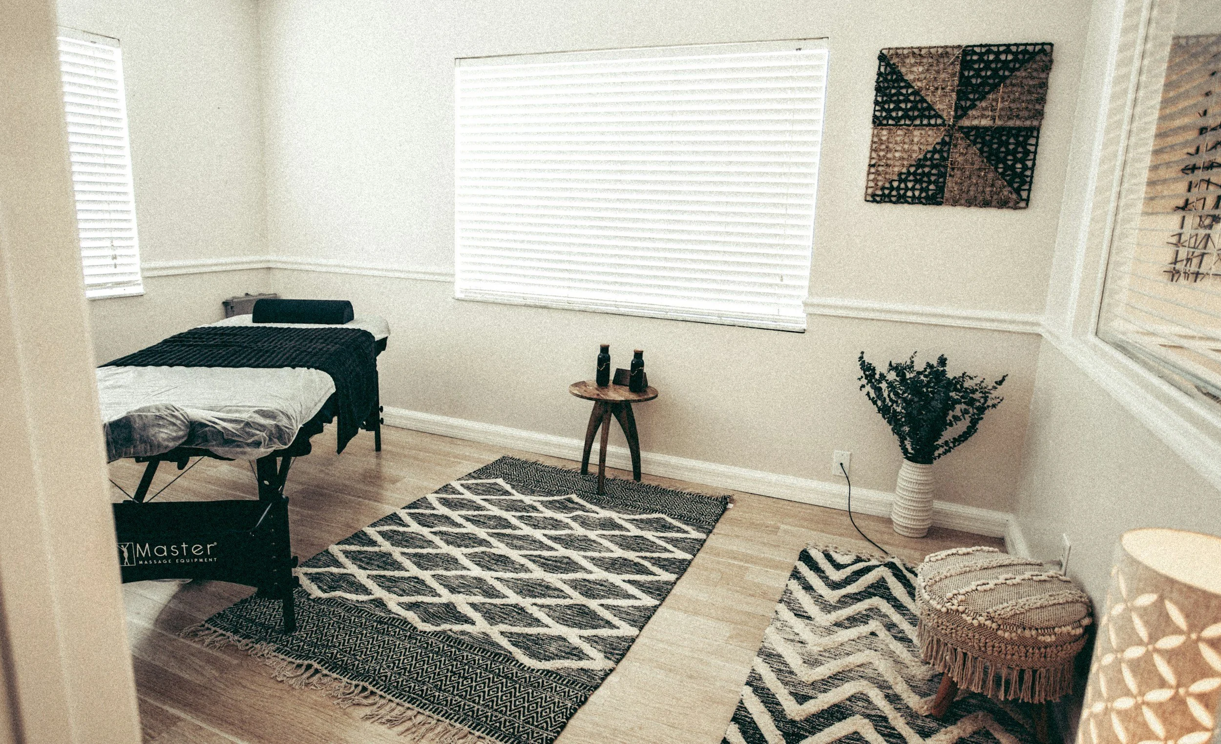 A massage therapy room with a massage table, black and white patterned rugs, a small wooden side table with three black decorative bottles, a large window with white blinds, and wall art. There is a leafy plant in a white vase and a small, round, woven stool.