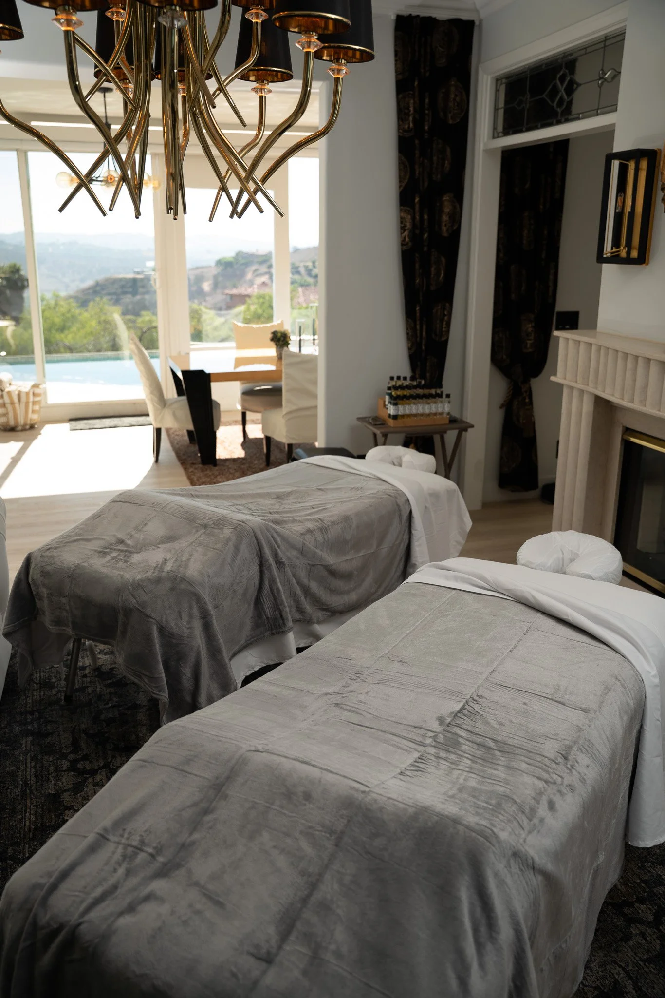 Two massage tables covered with gray towels in a well-lit room with large windows and a scenic view outside. There is a chandelier hanging from the ceiling.