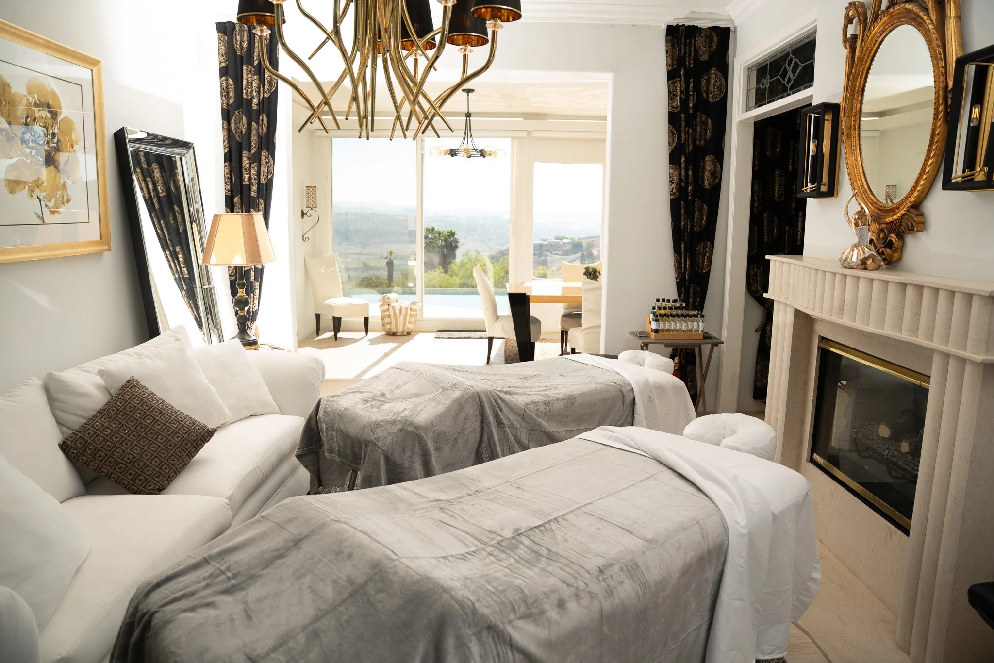 A luxurious room with two massage tables covered with gray throws and a white sofa with a patterned pillow. The room features a fireplace with a golden mirror above it and a large window with black curtains showing a scenic outdoor view with trees and hills.