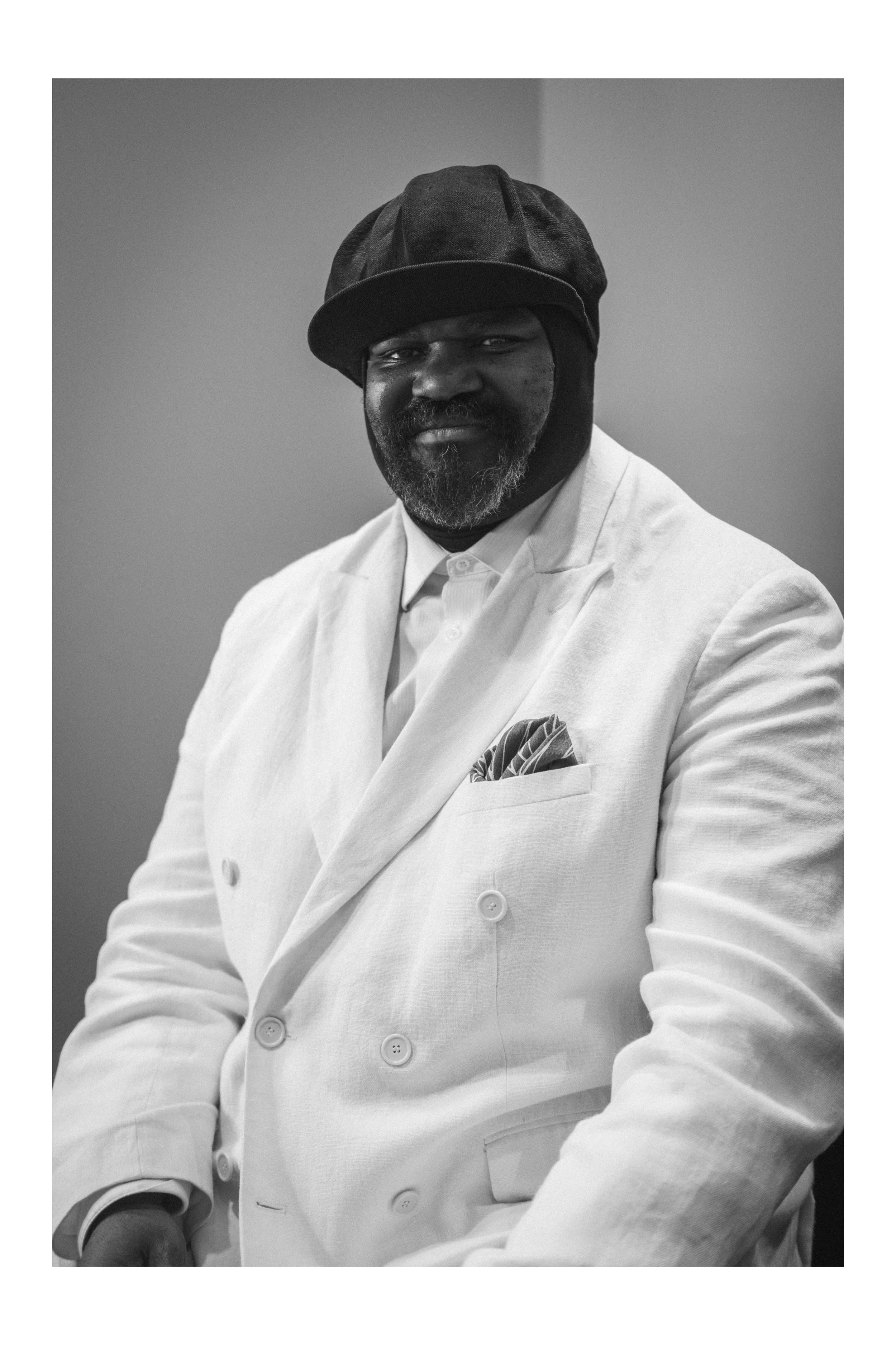 A man wearing a black hat, white suit, and pocket square poses for a black-and-white portrait.