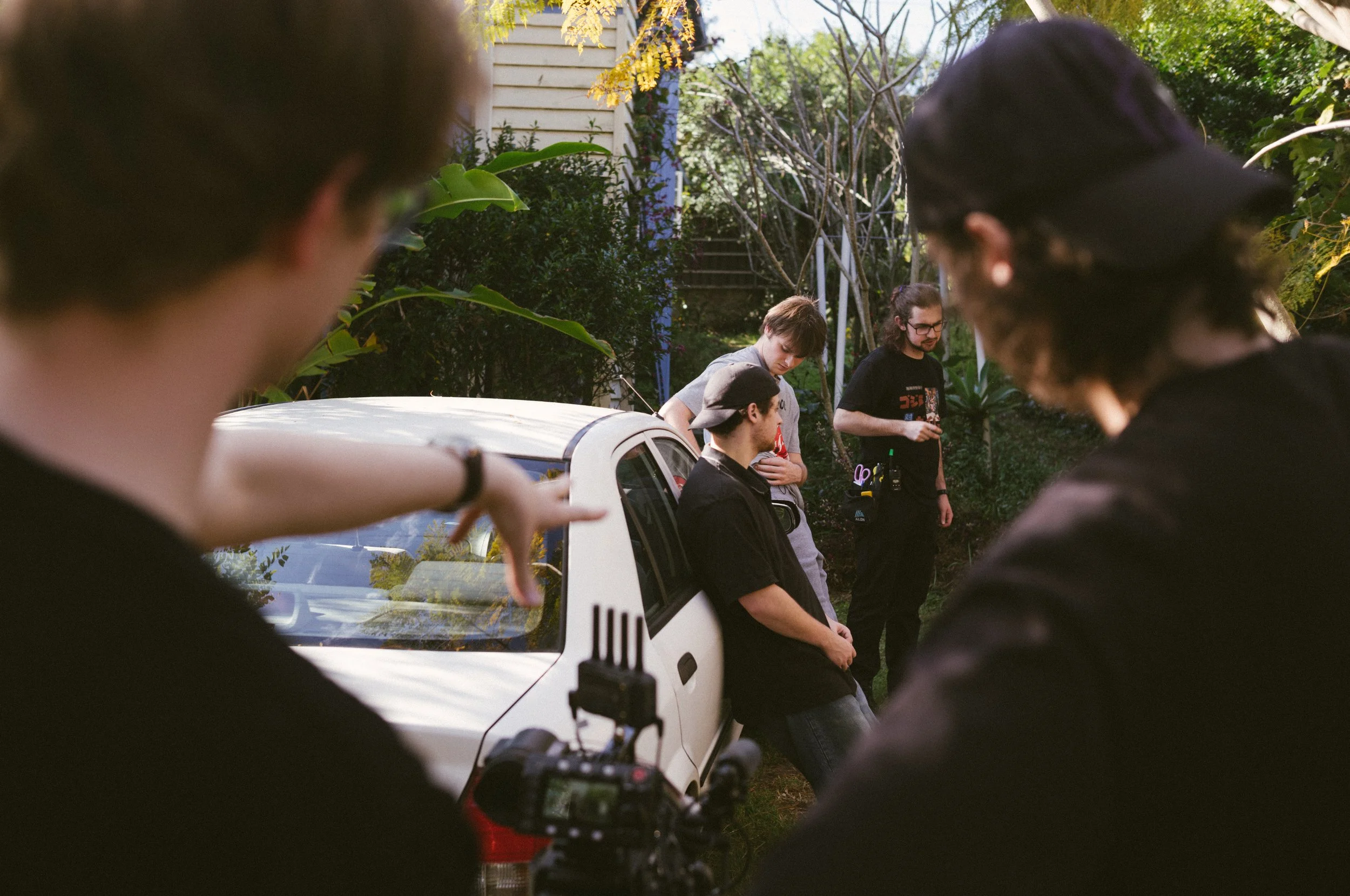People on a film set behind the scenes, with a white car and greenery in the background. One person is leaning on the car, with crew members behind, as a person points at the scene.