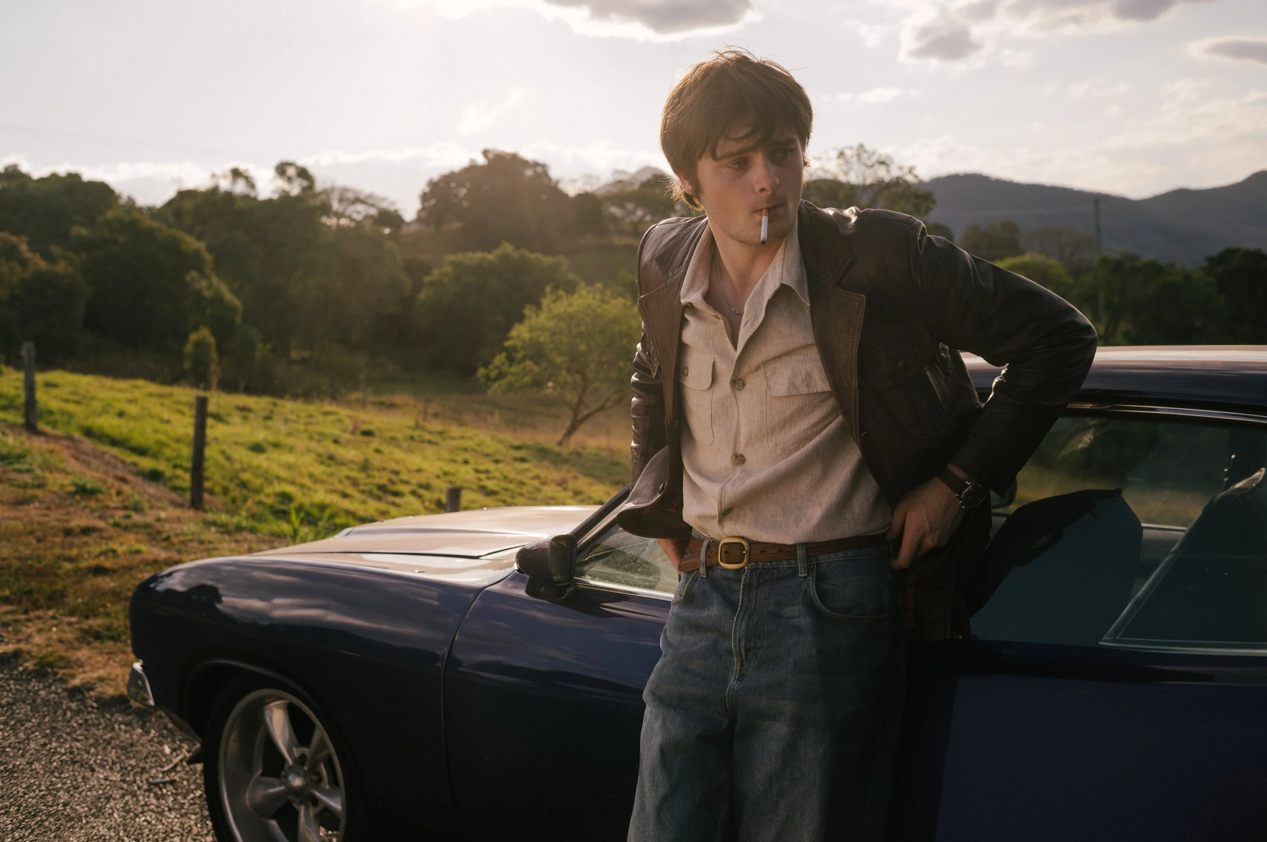 A young man with dark hair leaning against a classic black car, holding his right hand on his hip, with a cigarette in his mouth, wearing a beige shirt, dark leather jacket, and jeans, in a rural landscape with trees and mountains in the background d
