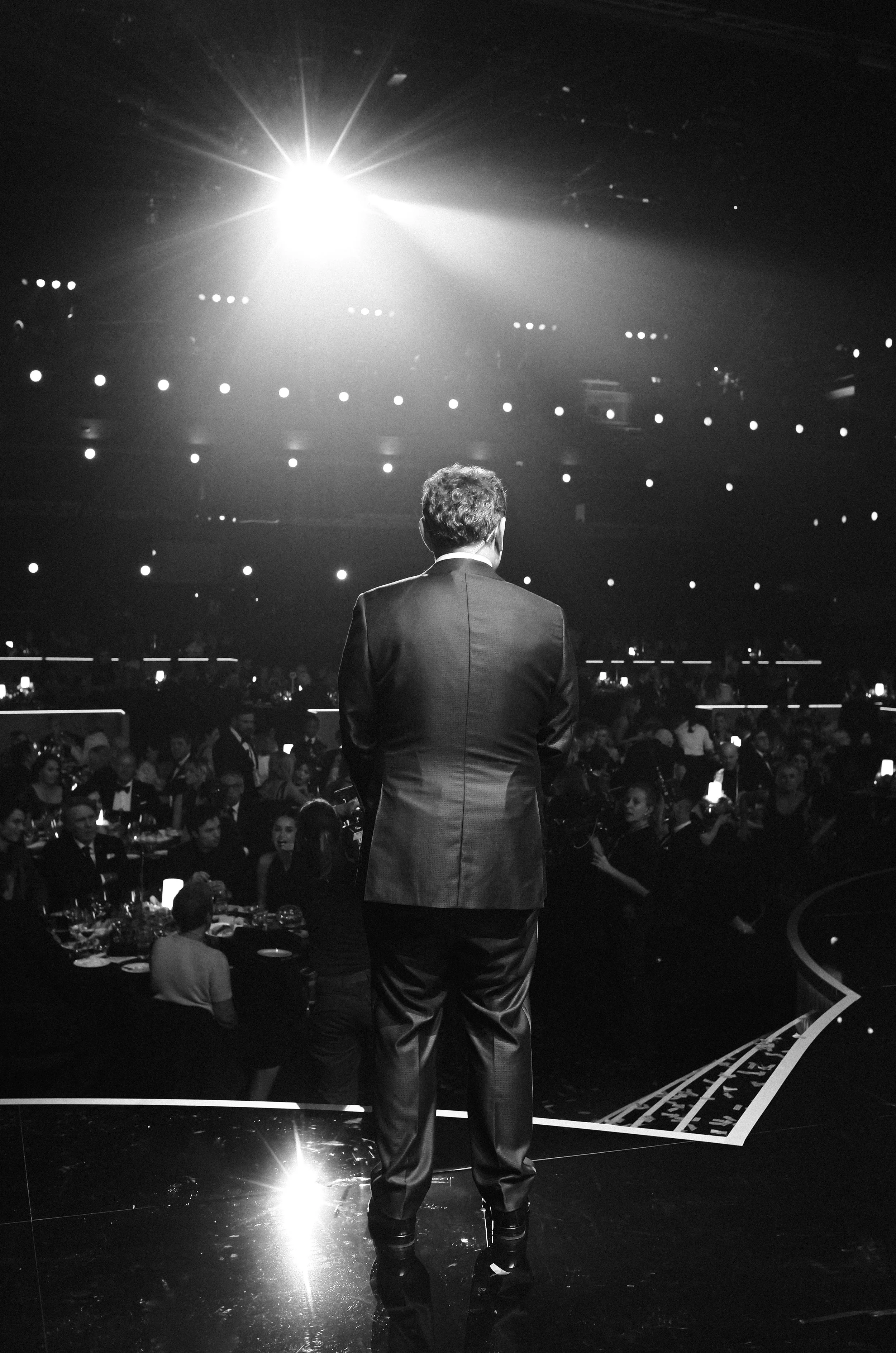 A man in a tuxedo standing on stage, facing an audience in a large, decorated auditorium with dramatic lighting.