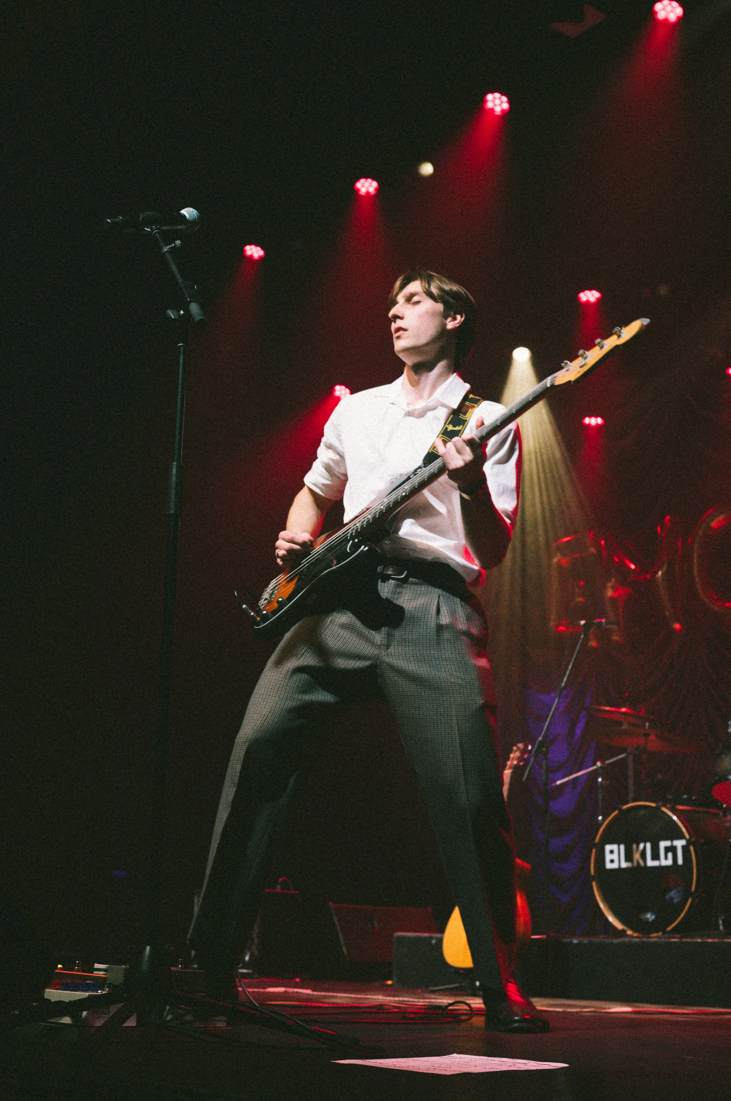 Male musician playing guitar on stage with red and yellow lighting, microphone stand nearby, drum set labeled 'BLK LGT' in background.