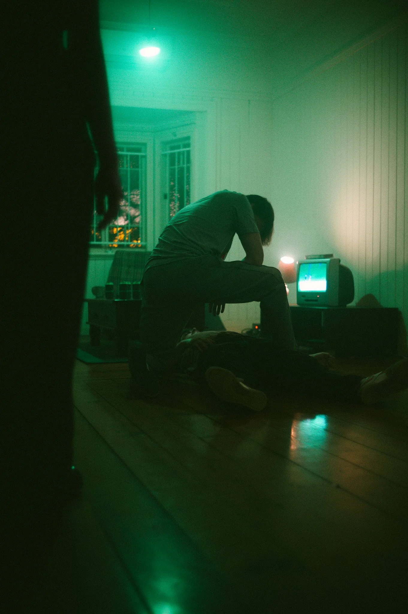 A person kneeling and leaning over another person lying on the floor, in a dimly lit room with green lighting, an old CRT television on a stand, and a window with bars.