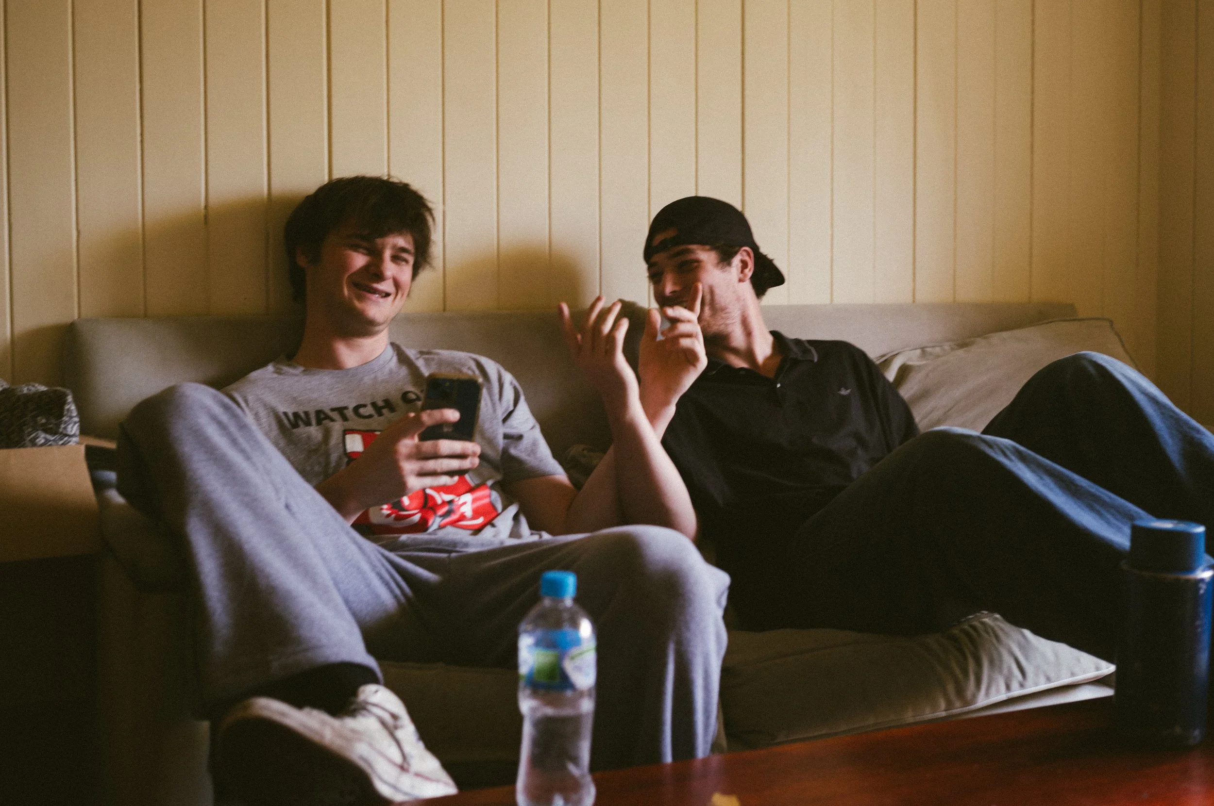Two young men sitting on a beige couch in a room with wooden panel walls. One is holding a smartphone and the other is gesturing with his hand. A water bottle is on the table in front of them.