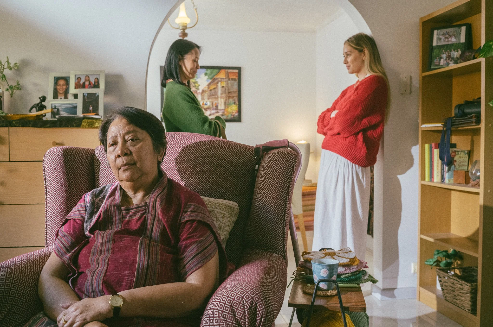 Three women in a living room. One woman is sitting in a chair with a neutral expression, the other two are standing and having a conversation.