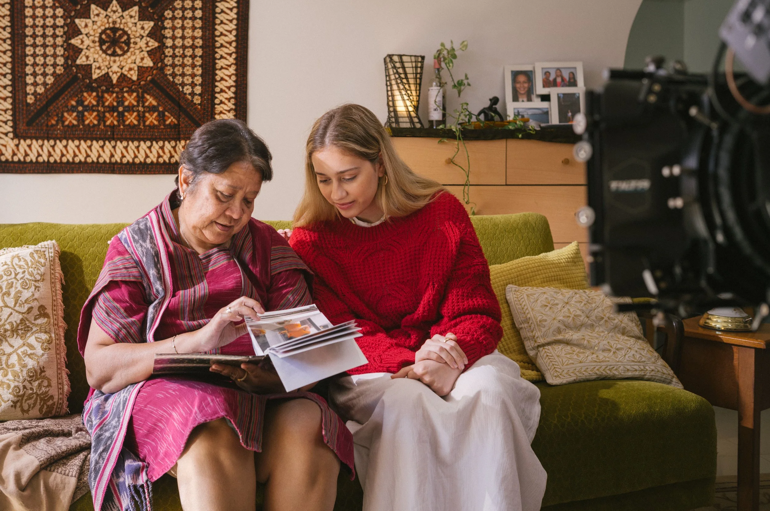 Two women, one older and one younger, sit on a green couch looking at a photo album together. The older woman wears a colorful, patterned dress and the younger woman wears a red sweater and white skirt. A TV camera is partially visible on the right.