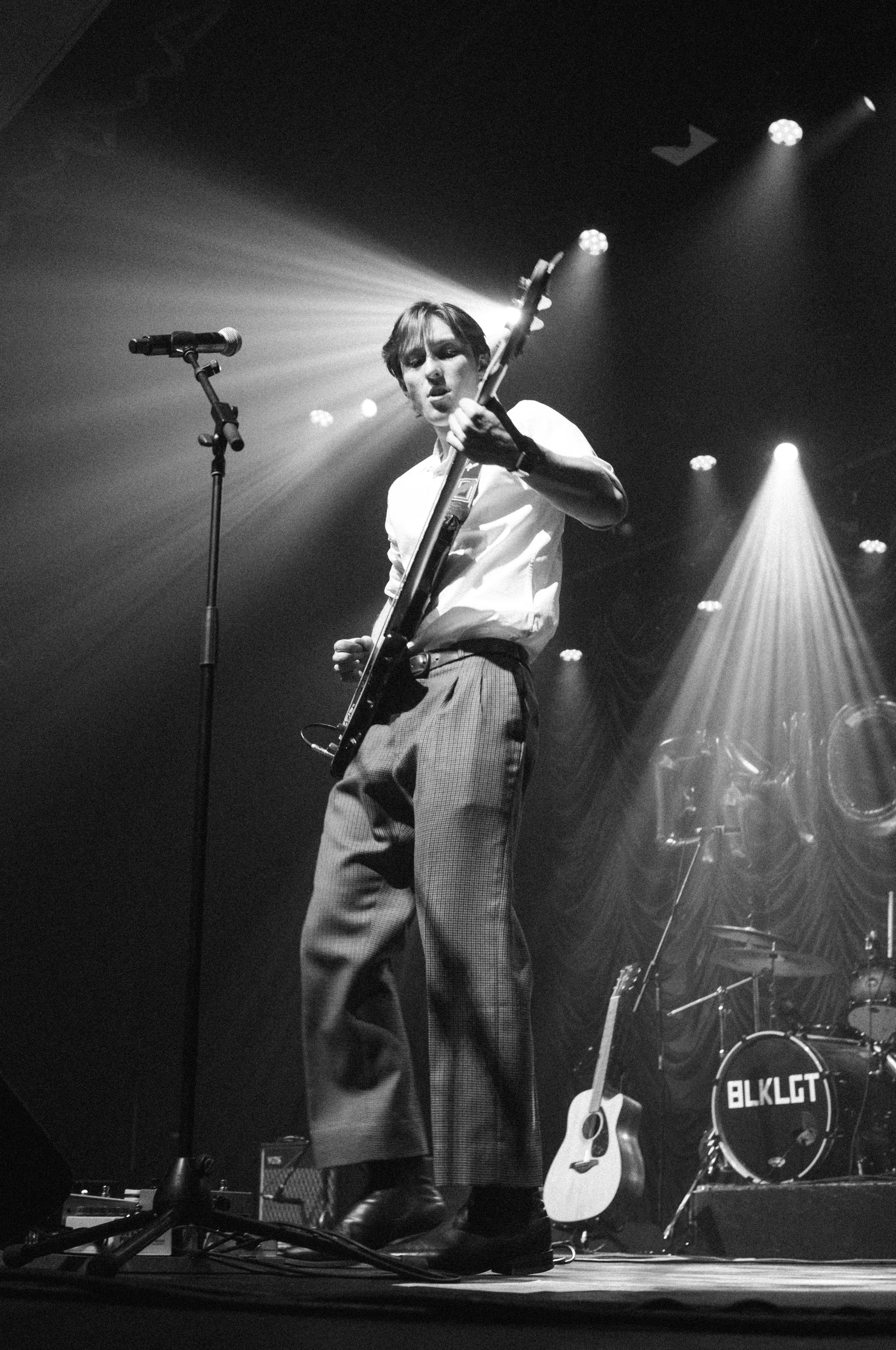A young man playing an electric guitar on stage during a concert, standing near a microphone with stage lights shining behind him.