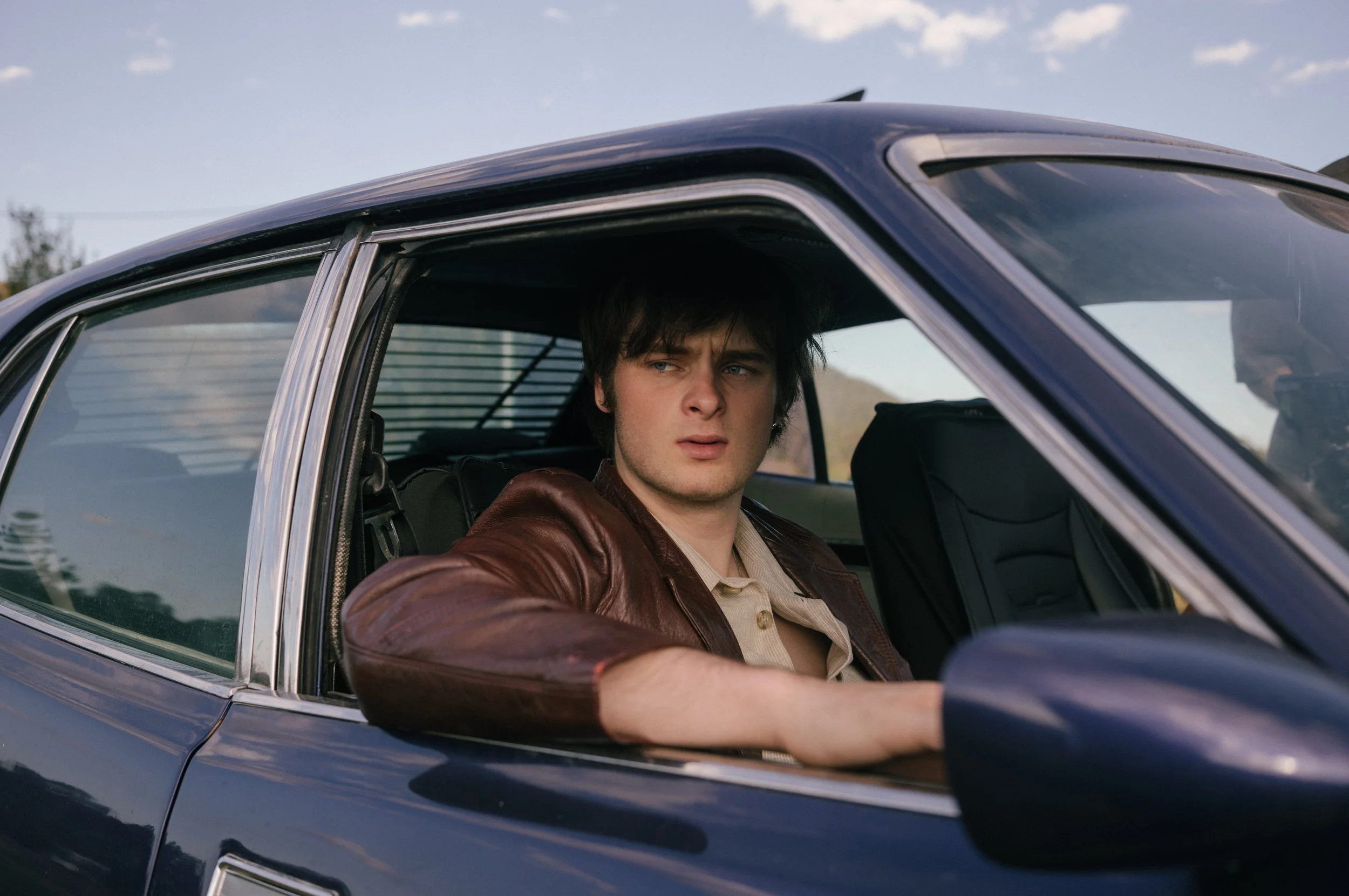 A young man with dark hair sitting in the driver's seat of a vintage blue car, looking outside with a serious expression.