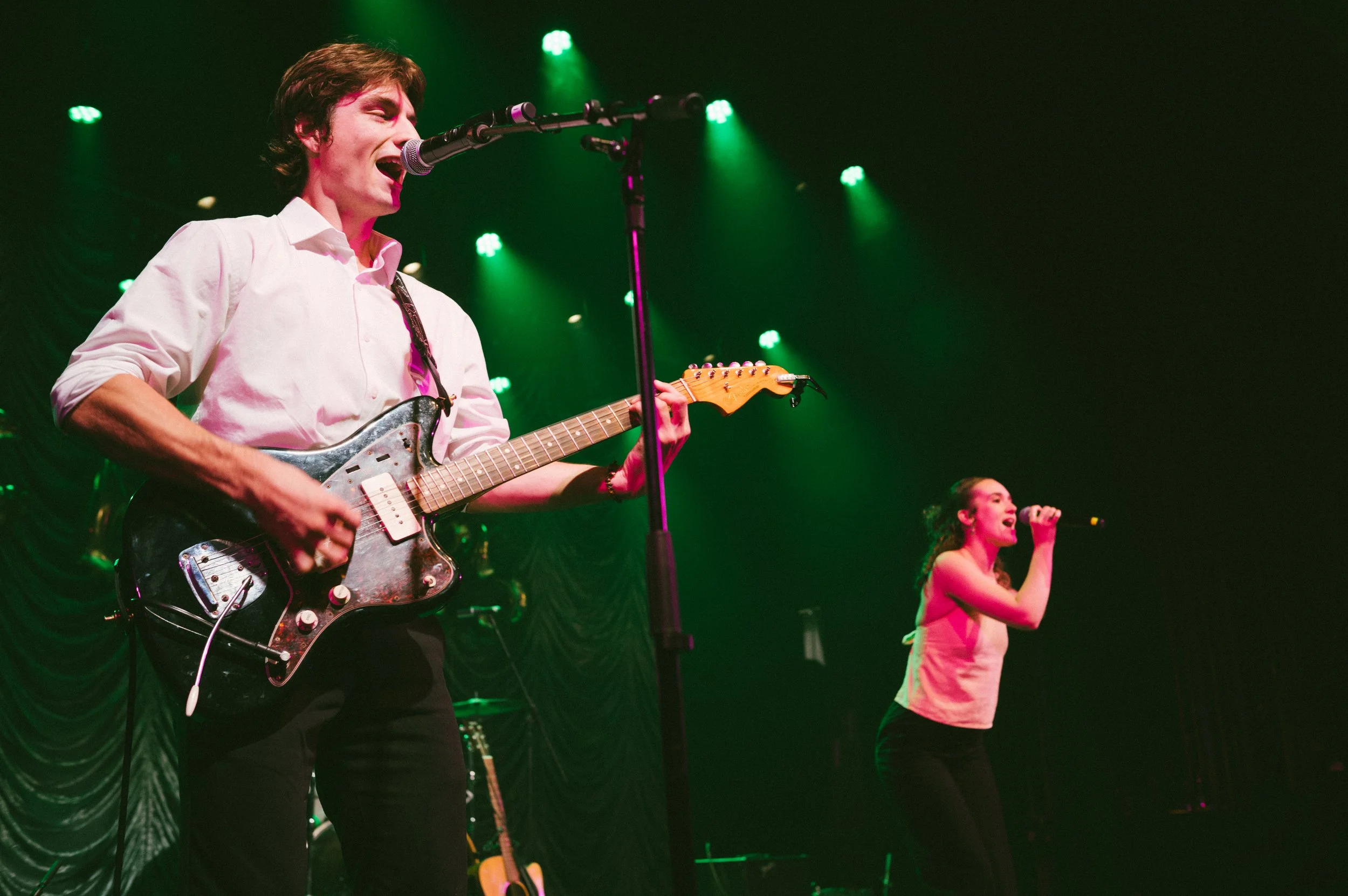 A man playing an electric guitar singing into a microphone on stage with a woman singing into a microphone in the background, with green stage lights 