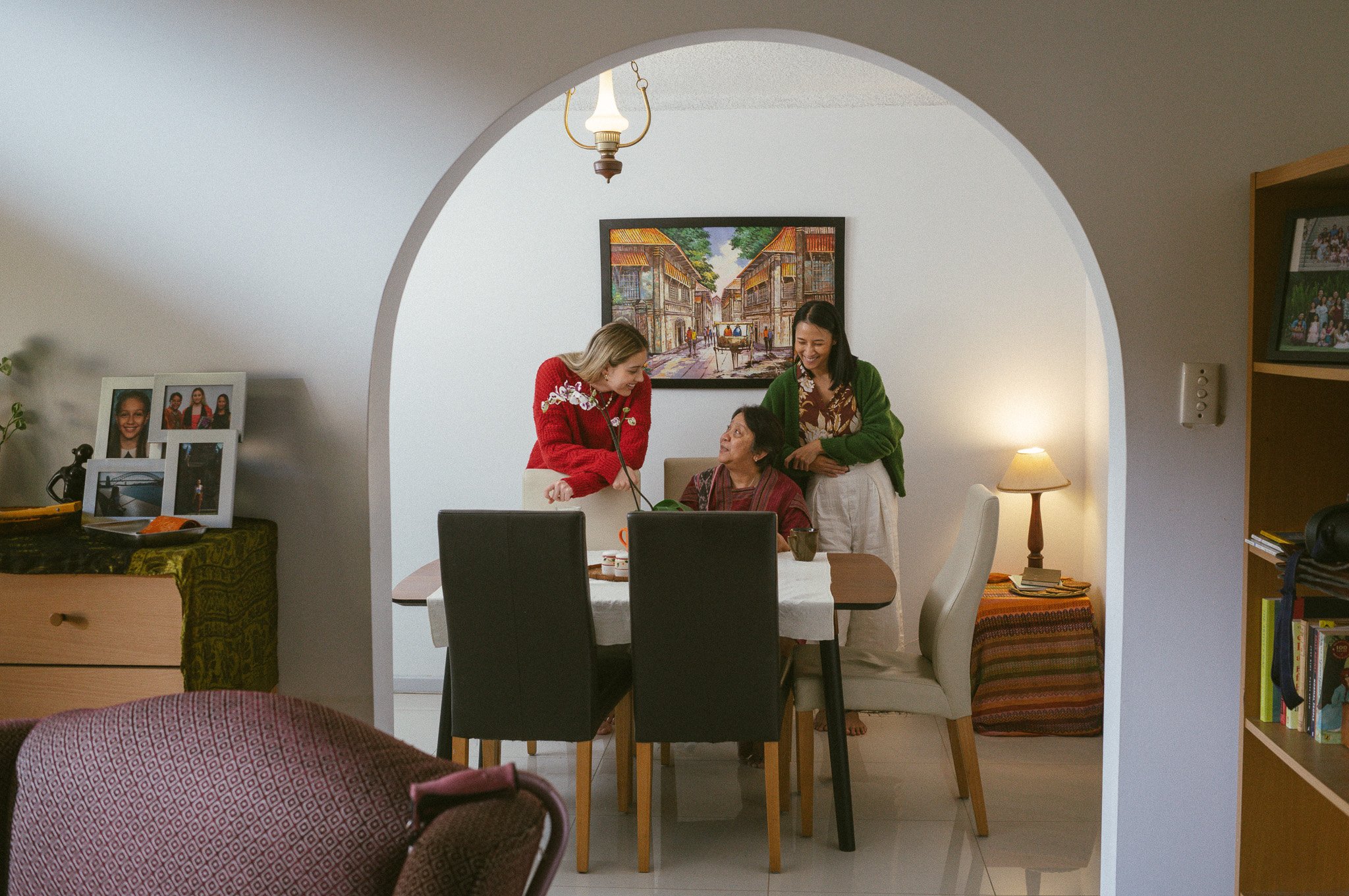 Three women gather around a dining table in a cozy home, smiling and talking, with a colorful painting on the wall behind them and a small lamp on a side table.