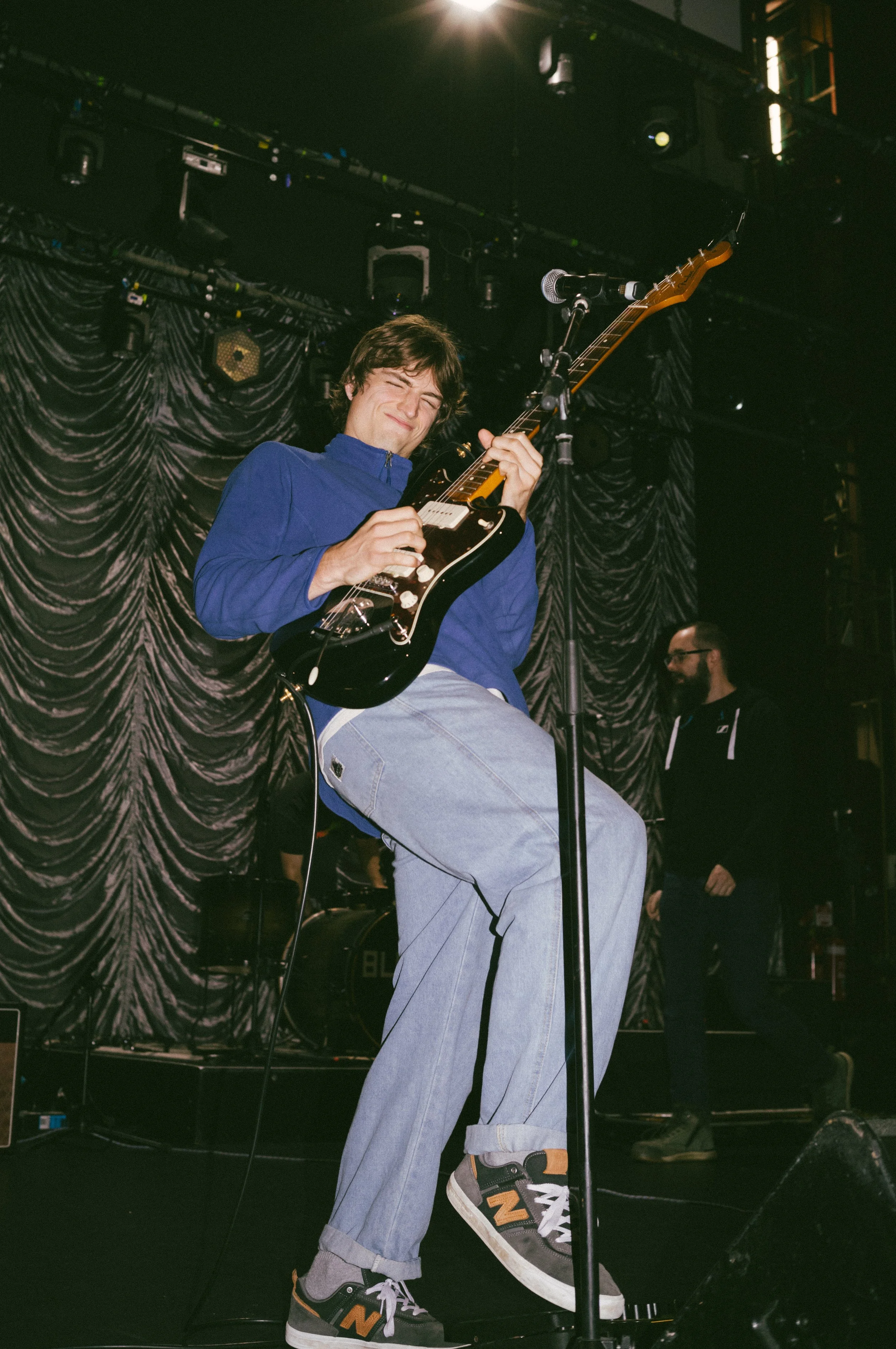 A young man playing an electric guitar on stage, wearing a blue jacket, light jeans, and sneakers, with a dark stage background and a person standing in the background.