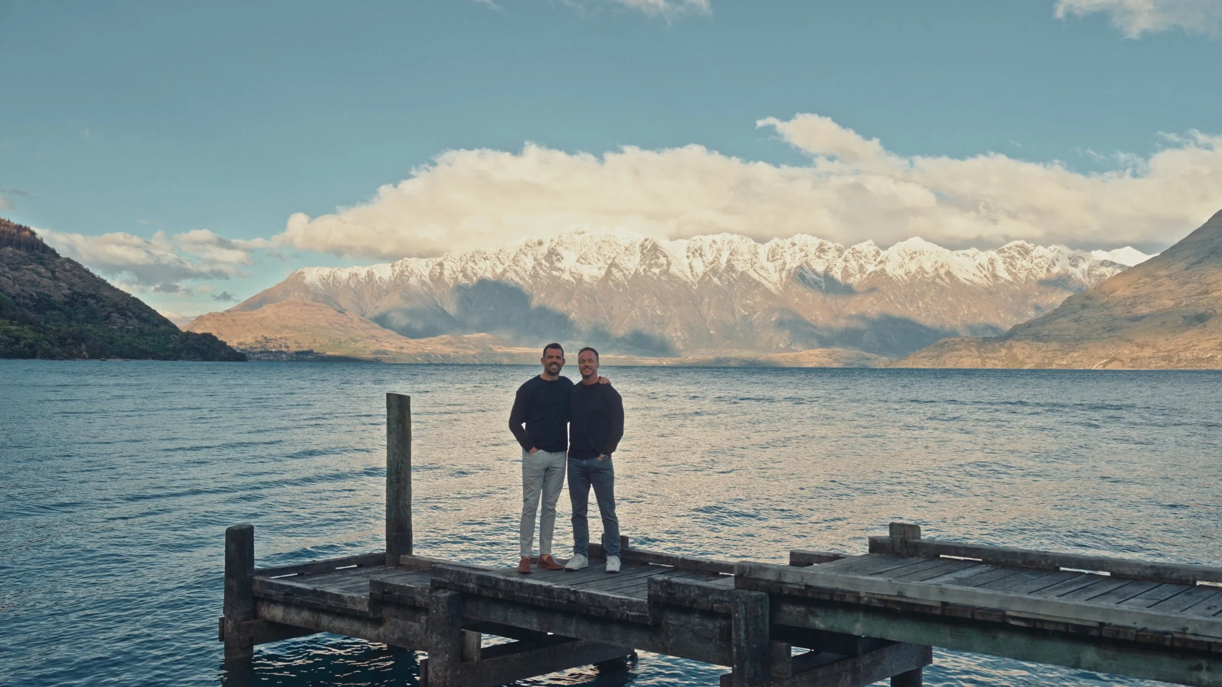Two men standing on a wooden dock by a lake with snow-capped mountains in the background.
