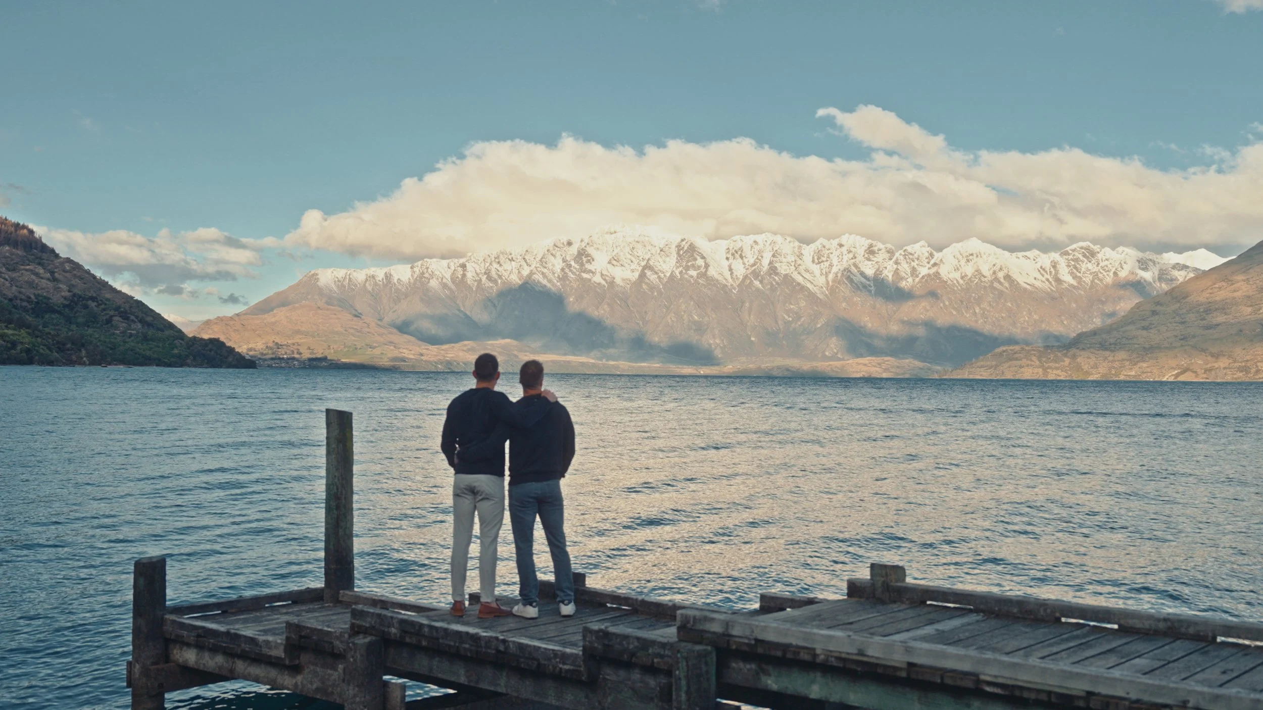 Two men standing on a wooden dock by a lake, with mountains in the background, some snow-capped peaks and cloudy sky.