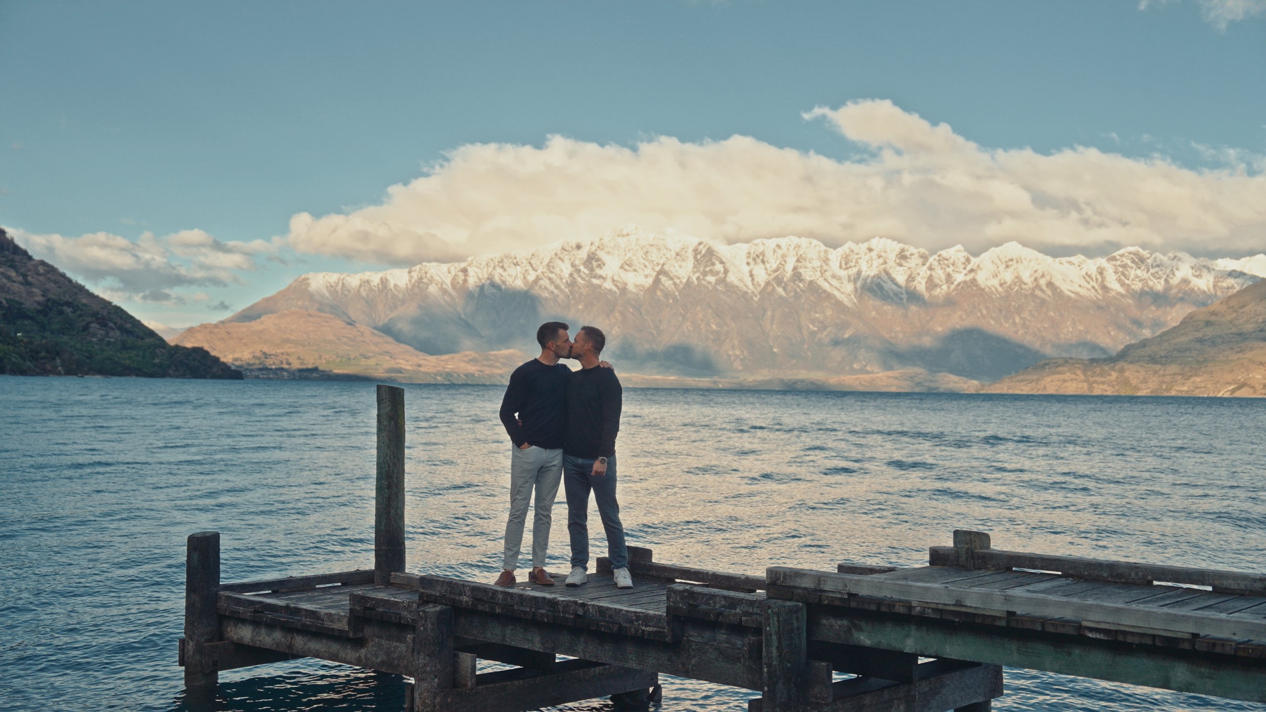 Two men kissing on a wooden dock by a lake with mountains in the background.