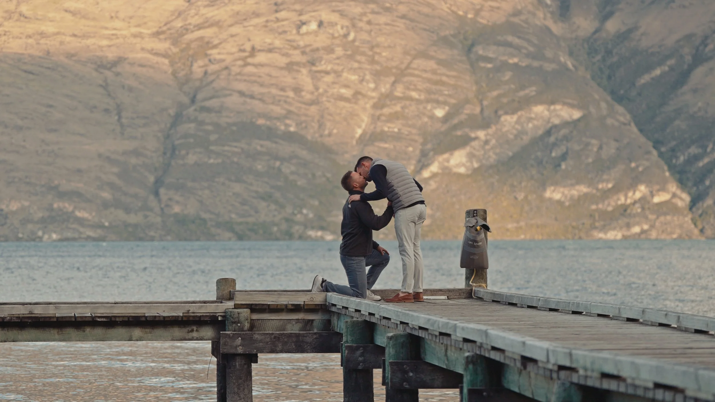 Two men on a wooden pier by a lake, one kneeling and proposing, the other standing and leaning in for a kiss, with mountains in the background.