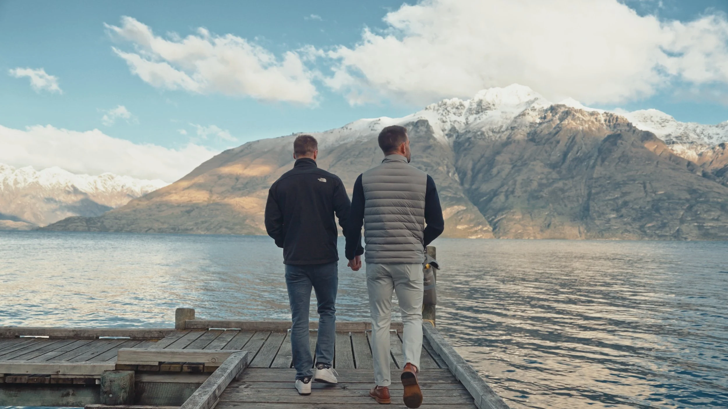 Two men walk hand in hand on a wooden dock overlooking a lake with snow-capped mountains in the background.