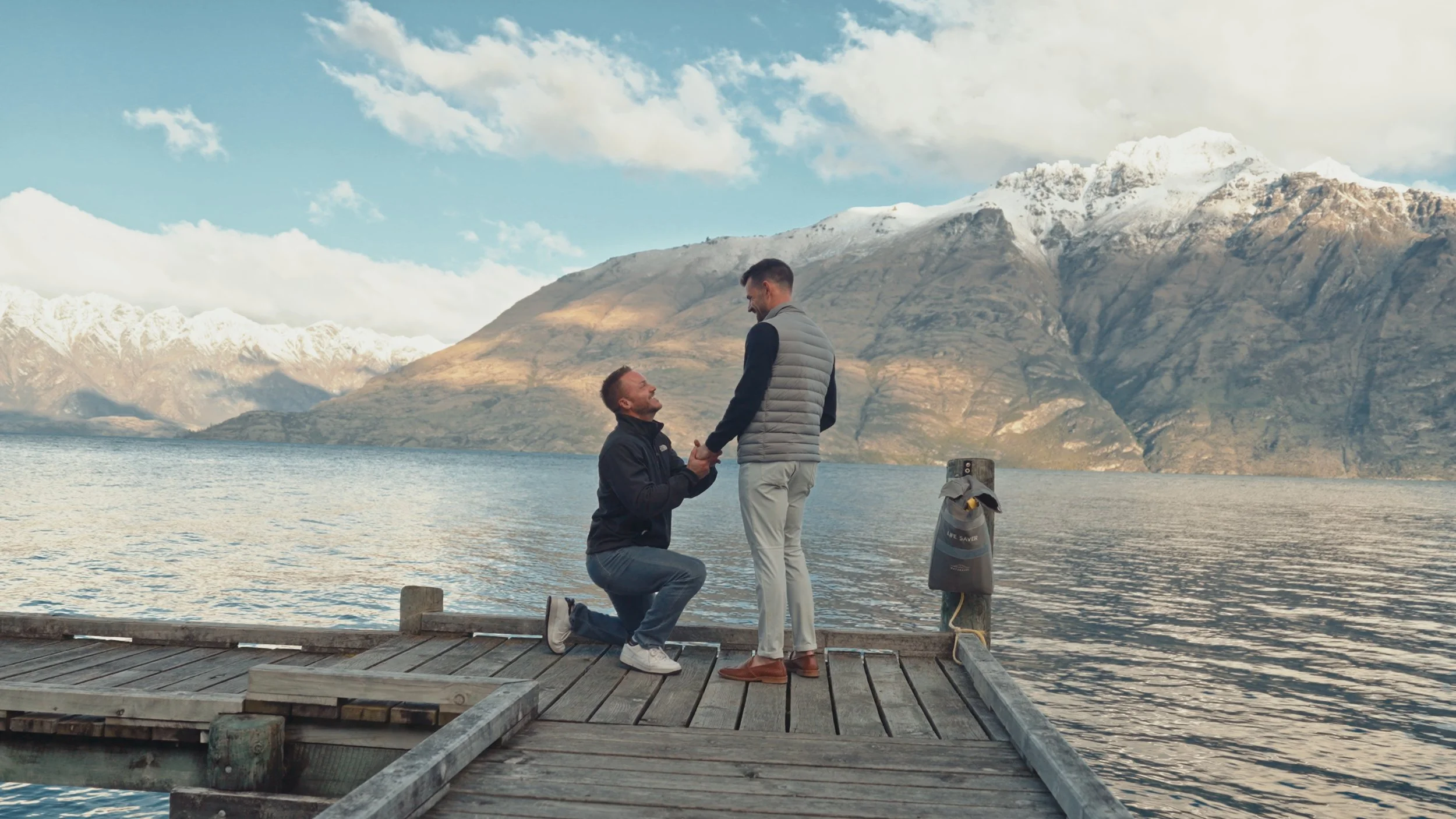 A man on one knee proposing to another man on a wooden dock by a lake with mountains in the background.