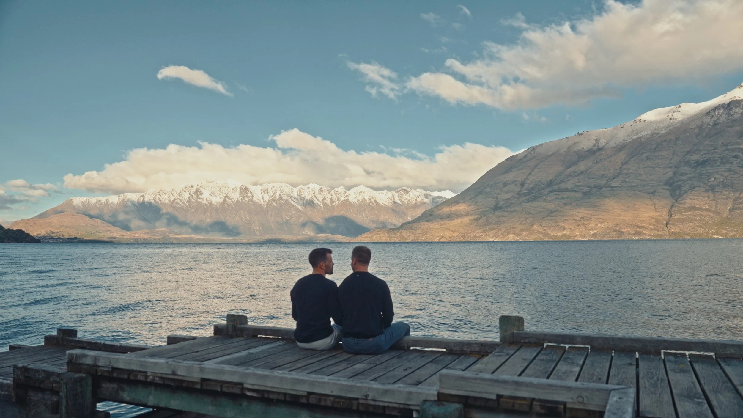 Two men sitting on a wooden dock by a lake, with snow-capped mountains and a partly cloudy sky in the background.