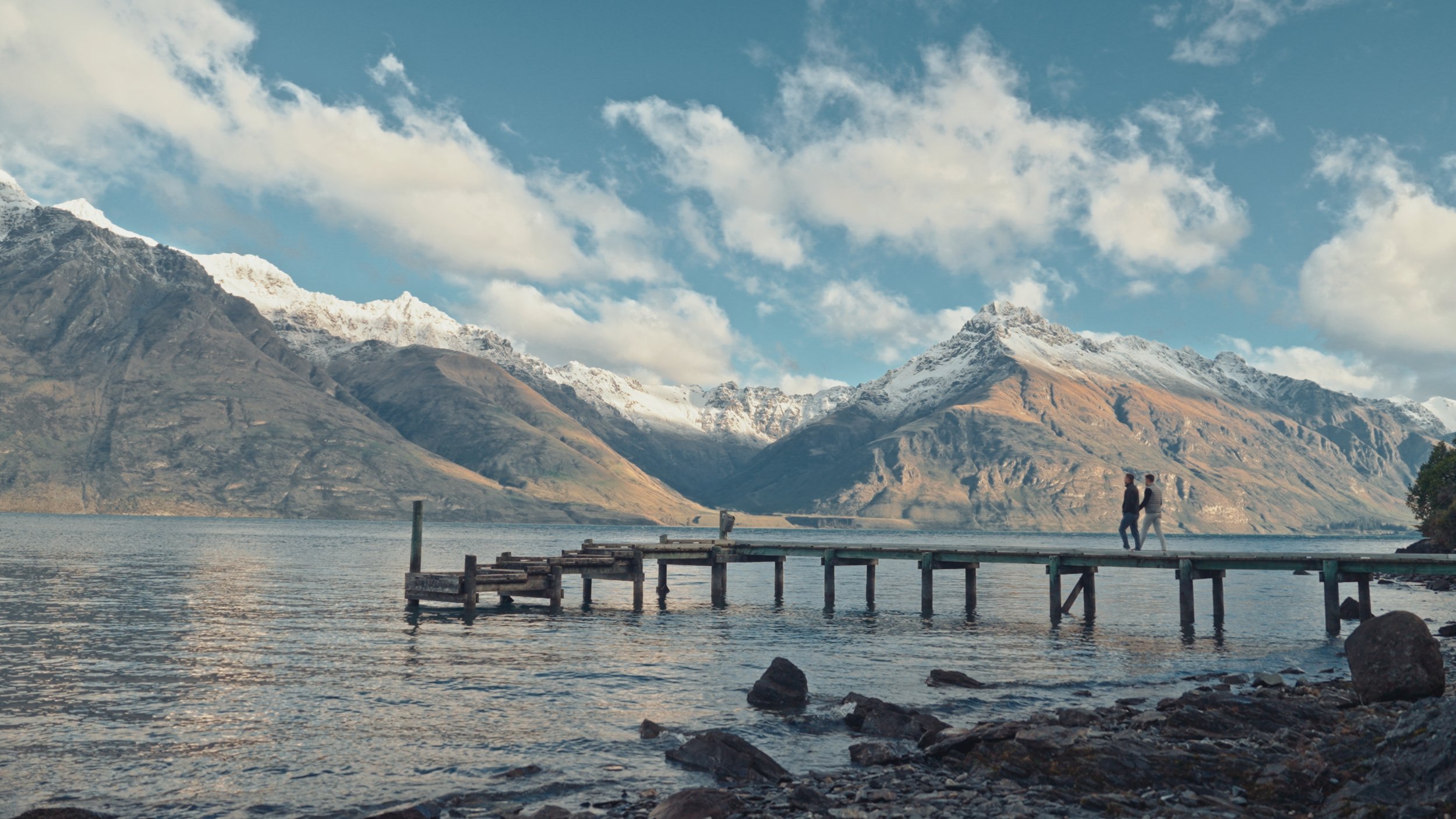 Two people walking on a wooden pier extending into a lake, with snow-capped mountains and a partly cloudy sky in the background.