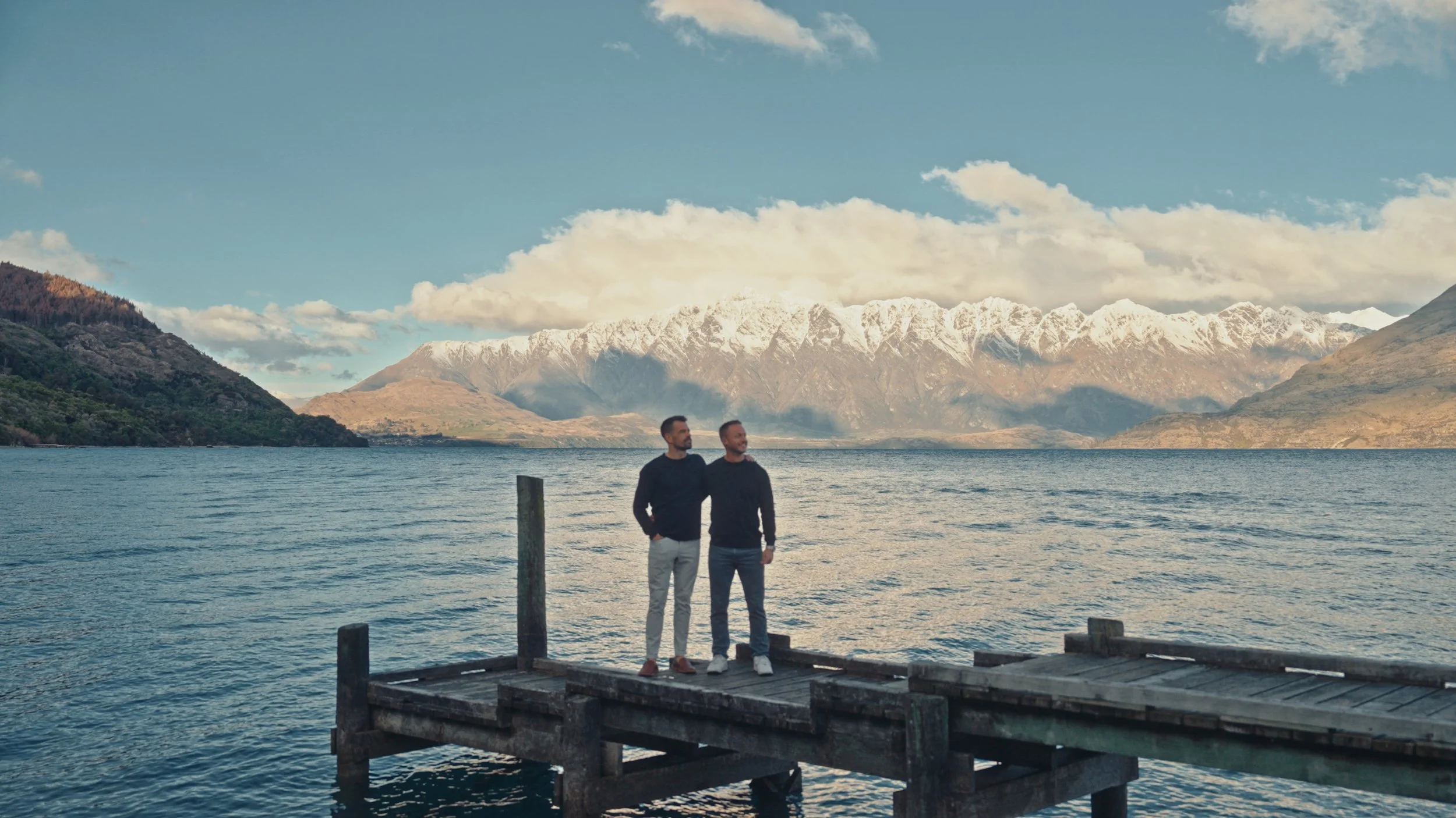 Two men stand on a wooden dock by a lake, surrounded by mountains with snow-capped peaks under a partly cloudy sky.