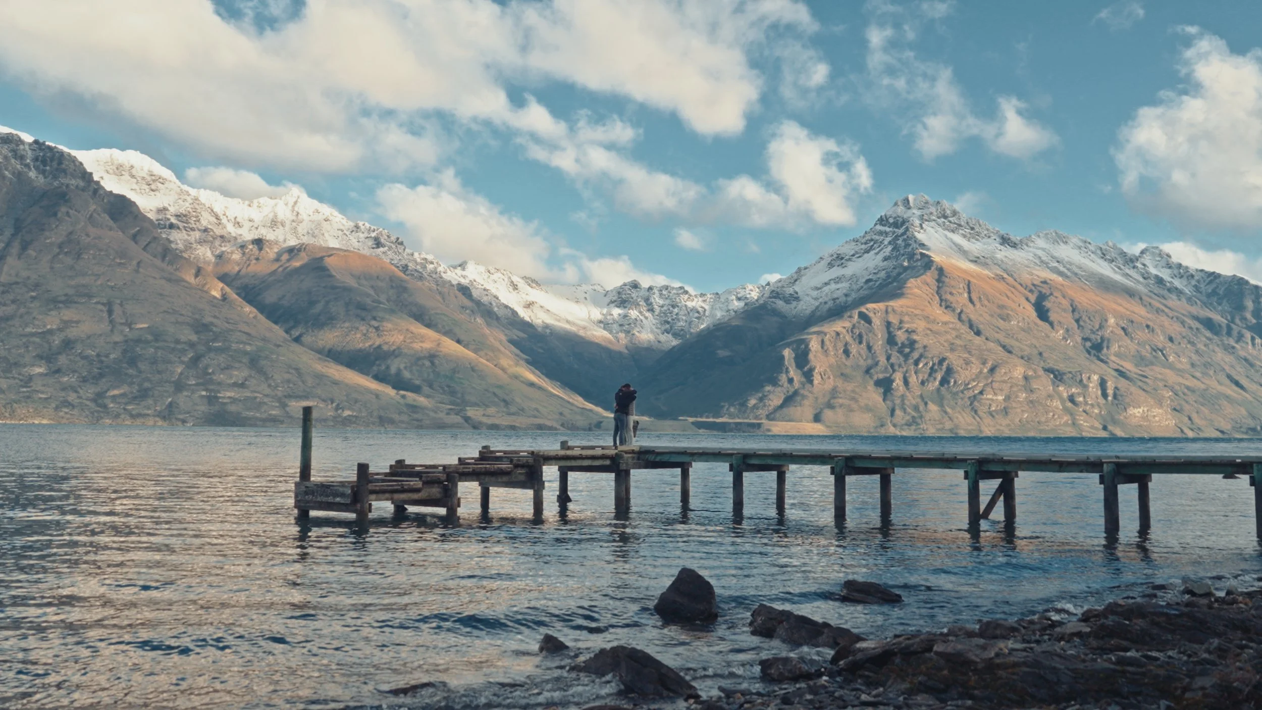 A person standing on a wooden pier at a lake, with snow-capped mountains in the background under a partly cloudy sky.