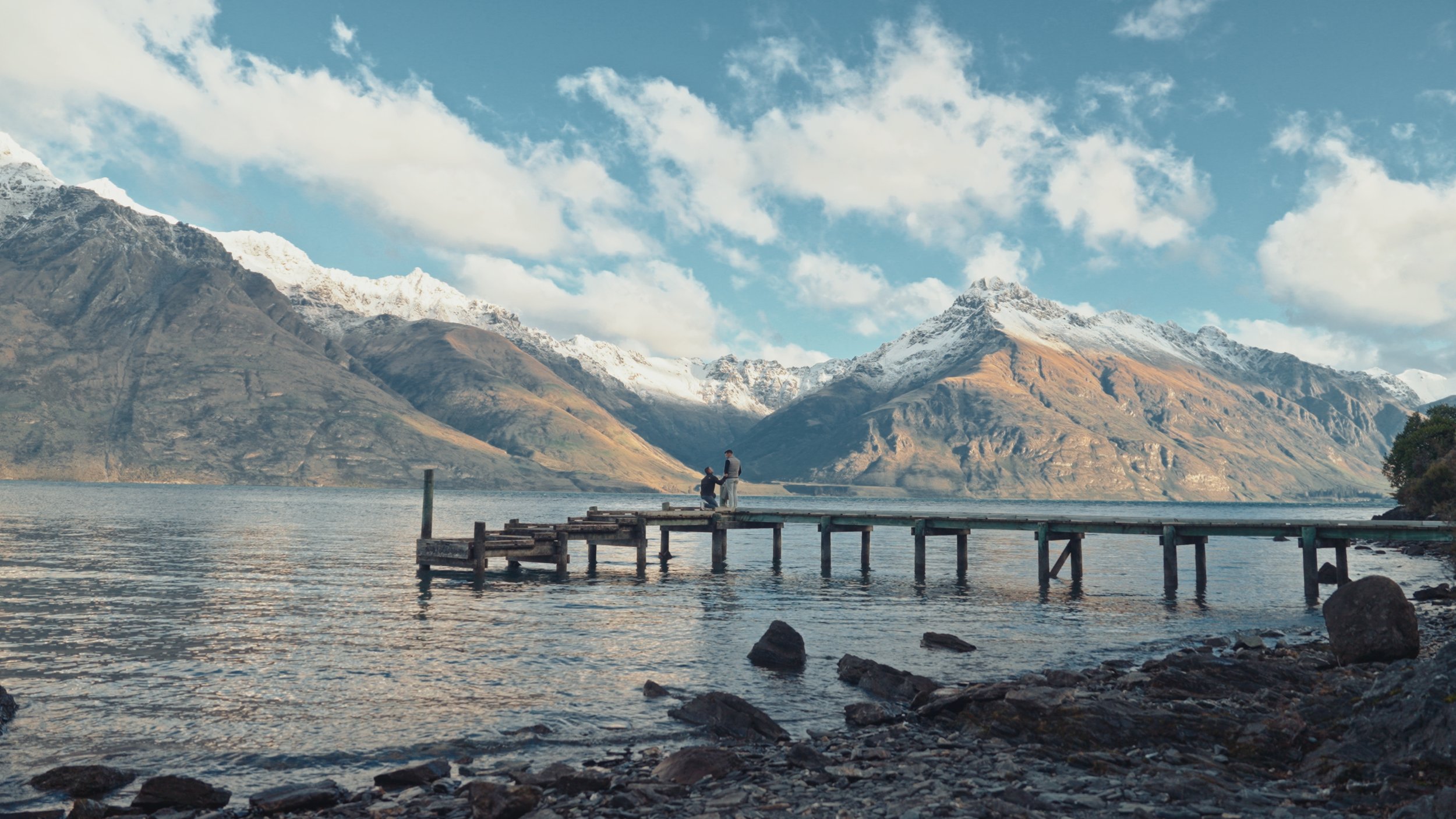 A dock extending into a lake with mountains and snow-capped peaks in the background, with two people standing on the dock.