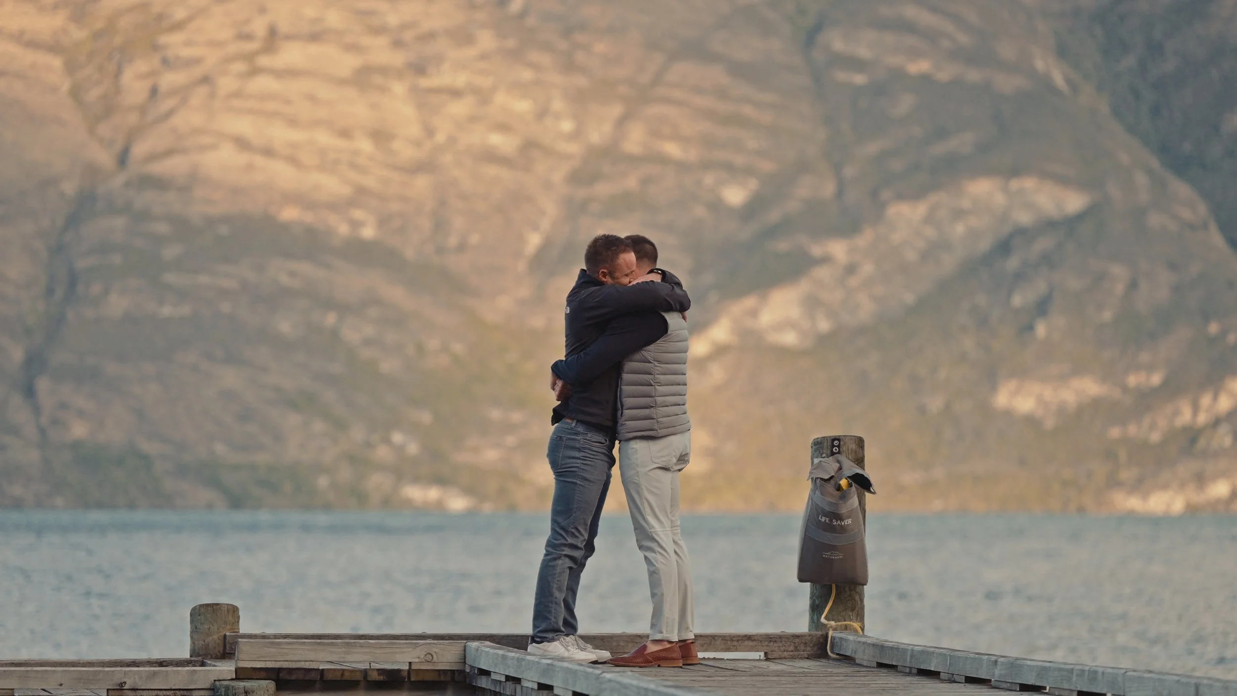 Two men hugging on a wooden dock by a body of water with mountains in the background.