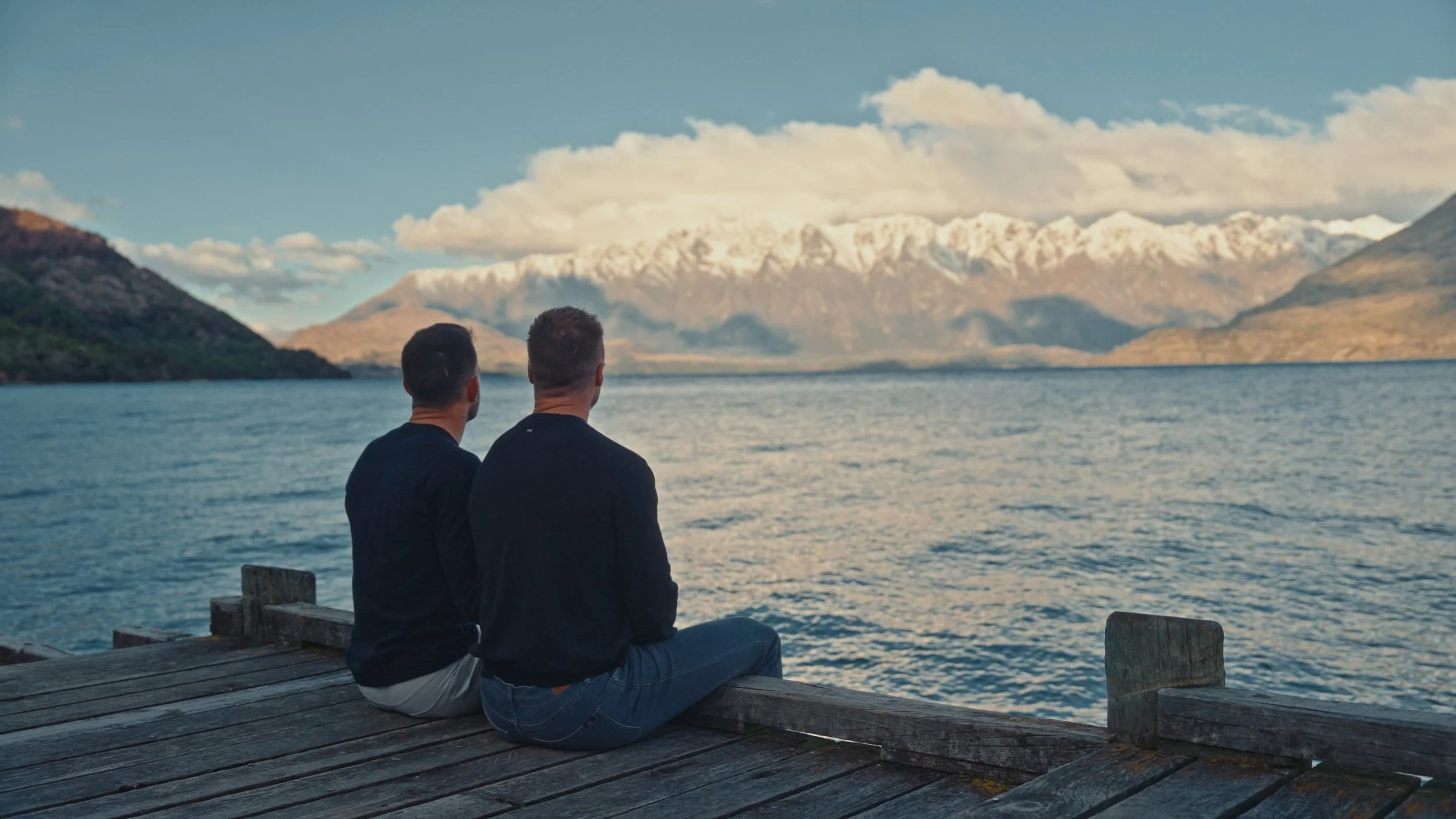 Two men sitting on a wooden dock overlooking a lake with snow-capped mountains in the background.
