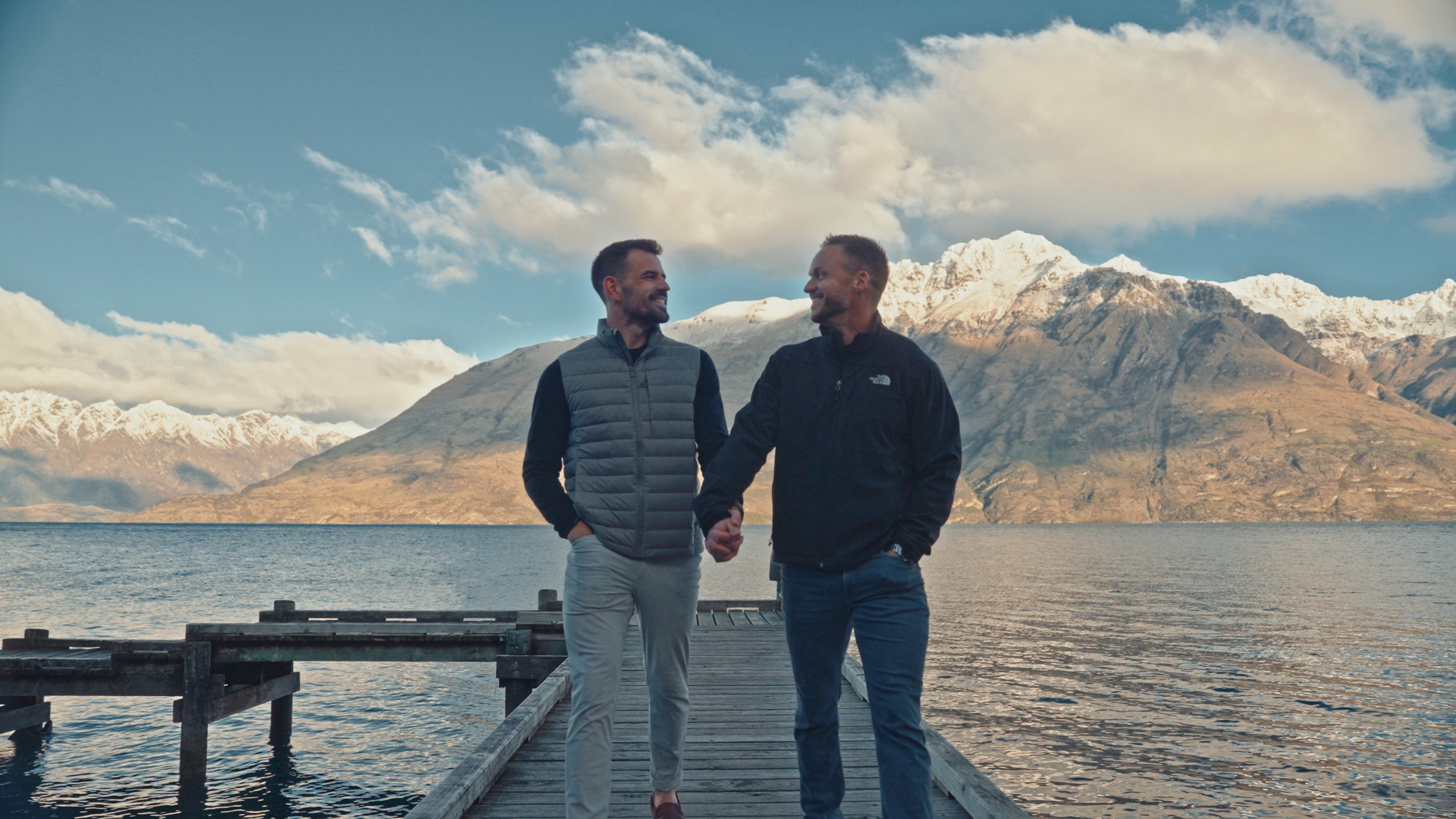 Two men shaking hands on a wooden dock by a lake with snow-capped mountains in the background under a partly cloudy sky.