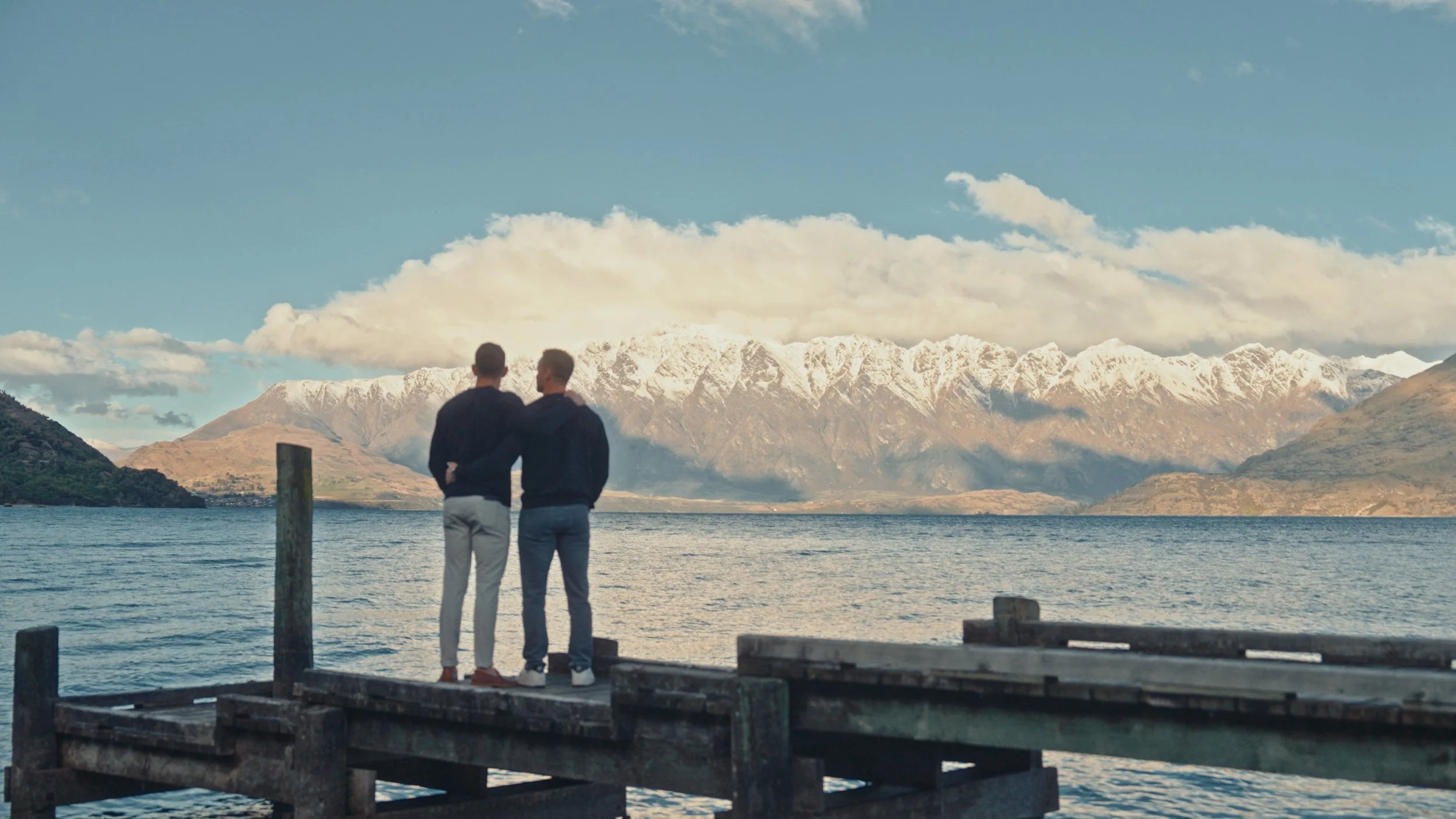 Two men standing on a wooden dock looking at snow-capped mountains across a lake.