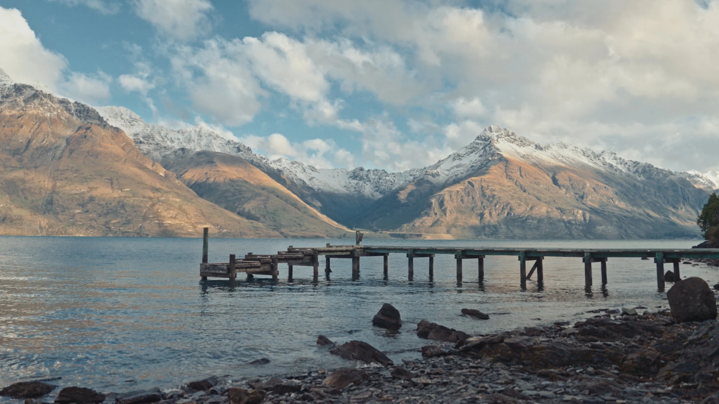 A wooden pier extends into a lake with snow-capped mountains in the background. The sky is partly cloudy.