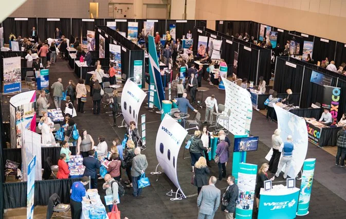 A busy trade show or conference with numerous booths and attendees walking around and engaging with displays.