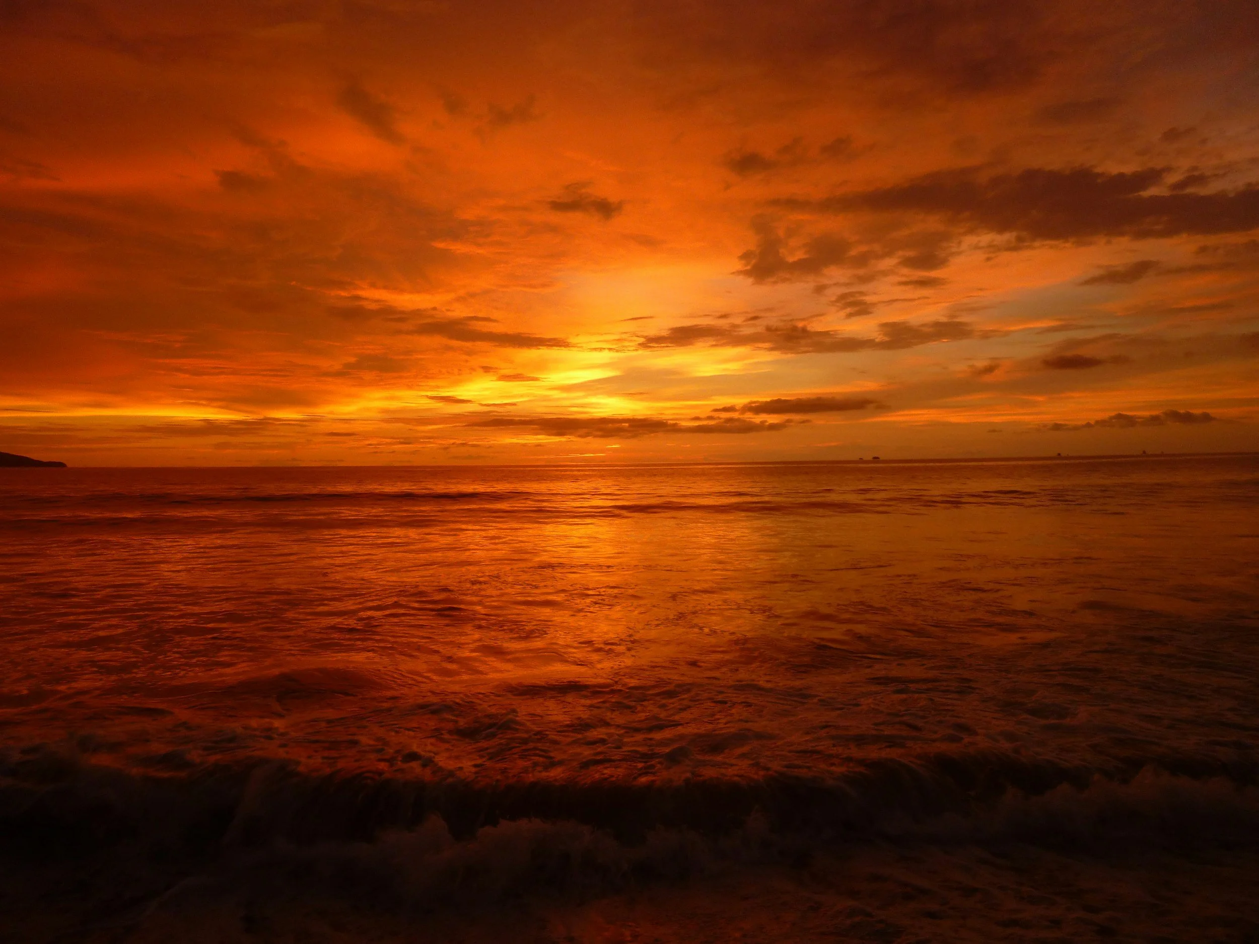 A vibrant sunset over the ocean with orange, yellow, and red hues reflecting on the water, clouds scattered across the sky, and a distant horizon.