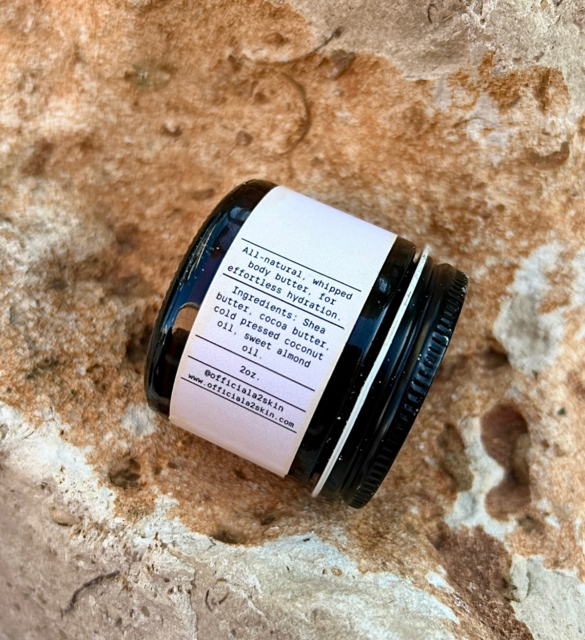 A small jar of whipped body butter with a white label, resting on a textured surface that appears to be sand or rough stone.