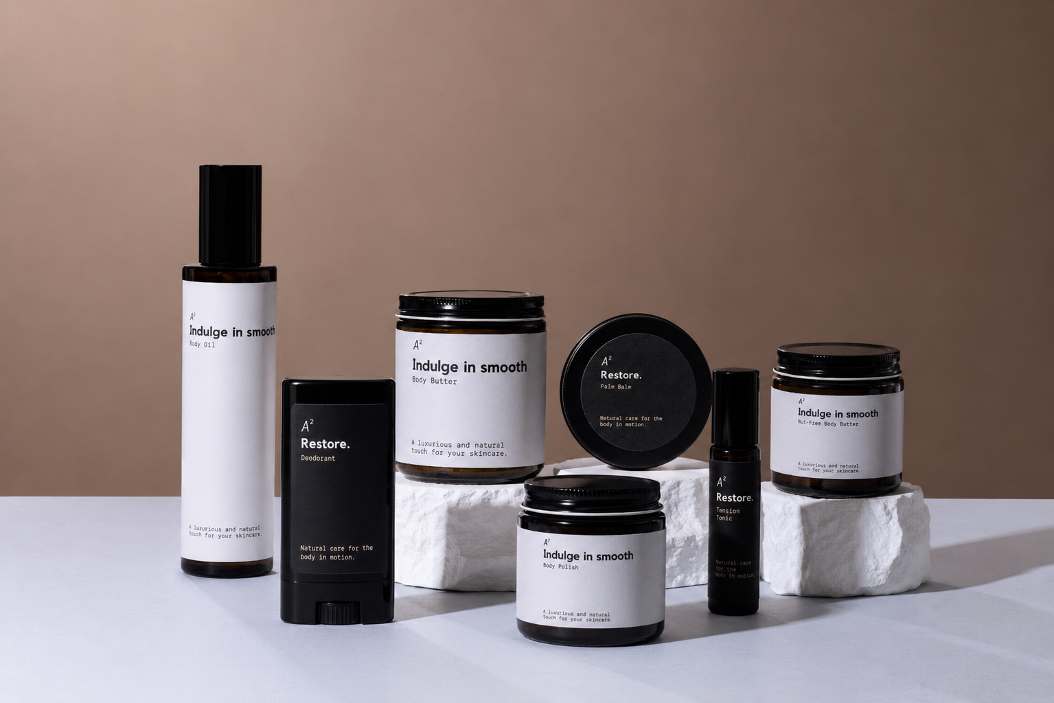 Collection of skincare products including body oil, body butter, body polish, deodorant, toner, natural, luxury skincare items with minimalist black and white labels arranged on white stone blocks against a neutral background.