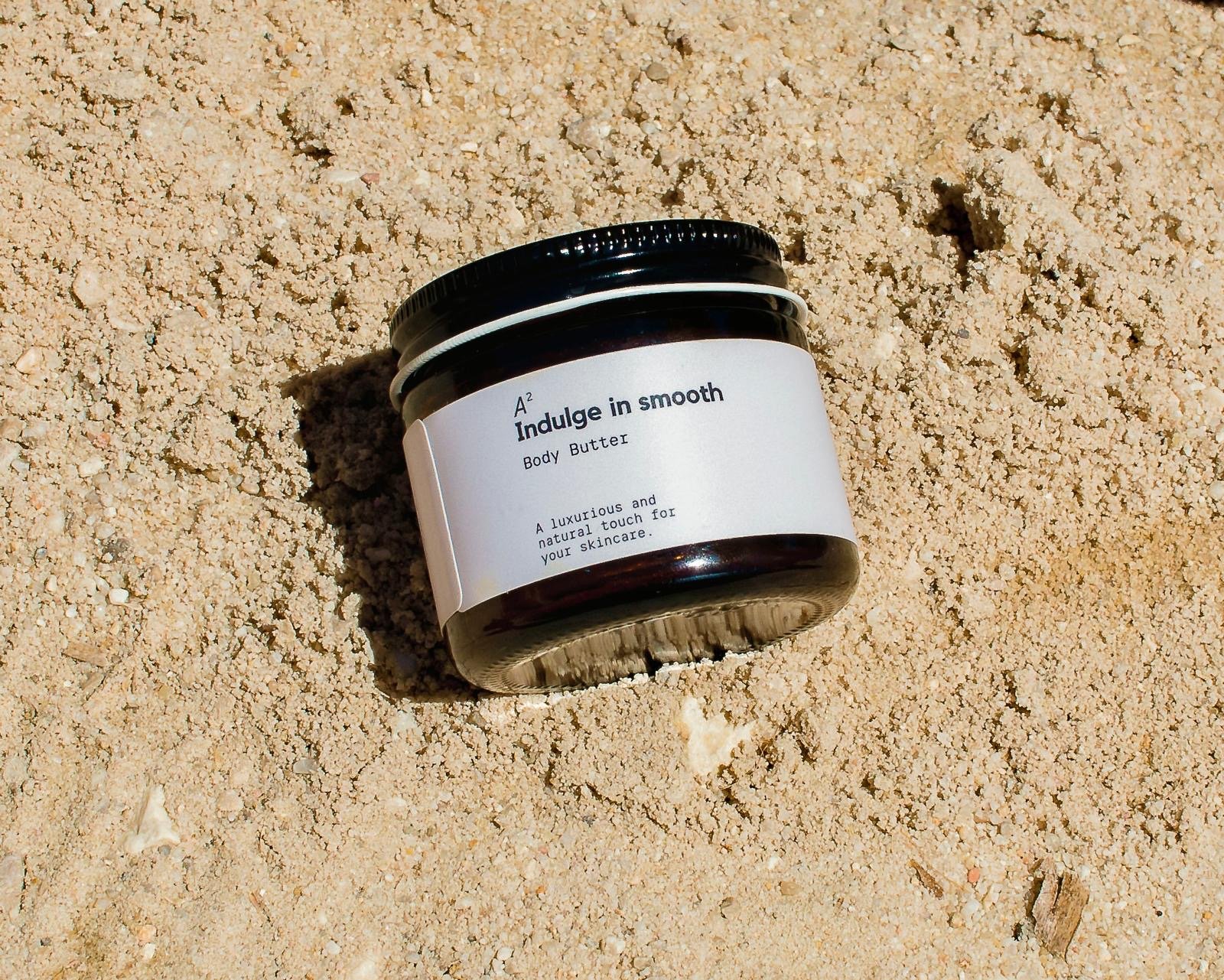 A jar of Indulge in smooth body butter lying on sandy ground, with the label visible.