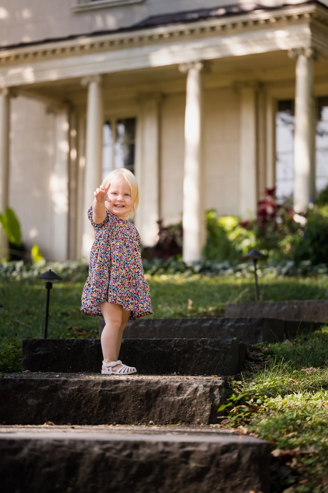 A young girl with blonde hair and a floral dress standing on outdoor steps, smiling and waving at the camera, with a house and garden in the background.