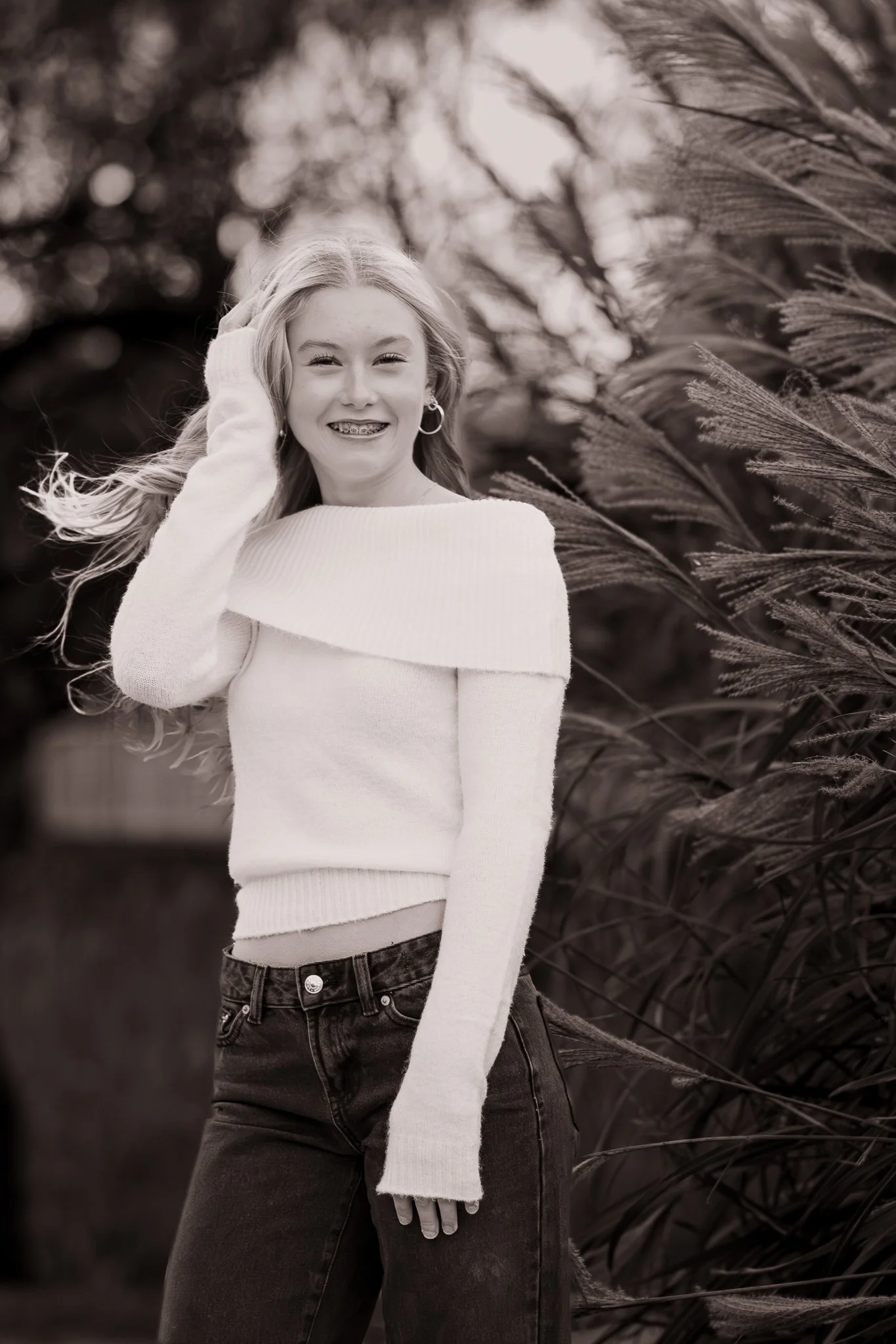 A young woman with long hair, wearing a white off-the-shoulder sweater and dark jeans, is smiling outdoors near tall grass Nancy Smiley Photography.