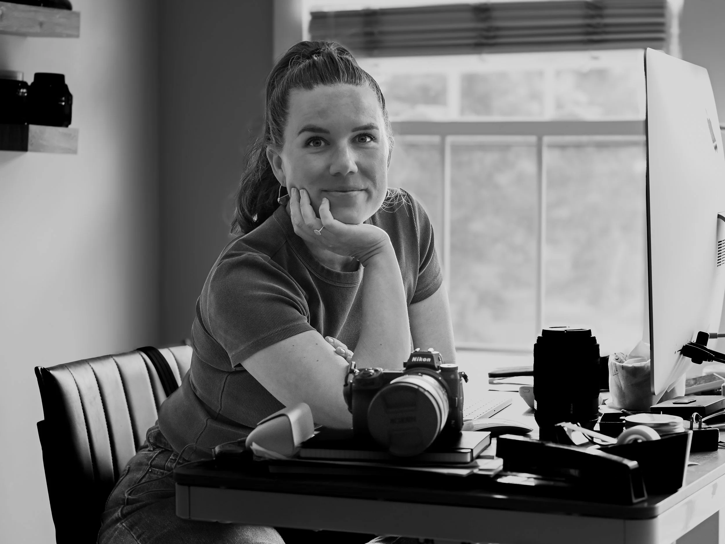 A woman with her hair tied back sits at a desk with a computer, camera, and camera lens, resting her chin on her hand, looking at the camera, in a well-lit room with a window in the background.
