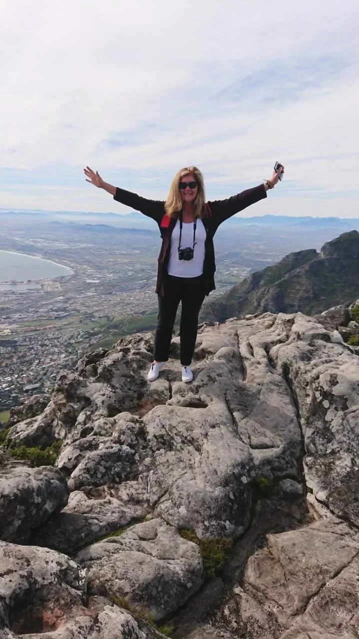 Woman standing on rocky mountain top with arms outstretched, wearing sunglasses, with a scenic view of a city, ocean, and mountains in the background.
