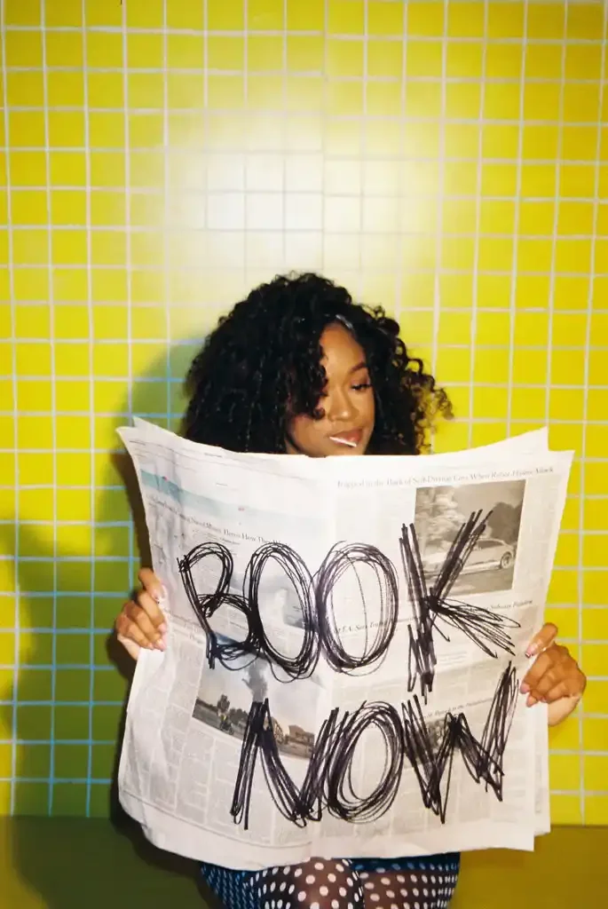 A young woman with curly hair is sitting against a bright yellow tiled wall, reading a newspaper with the words 'BOOK NOW!' written on it in black marker.