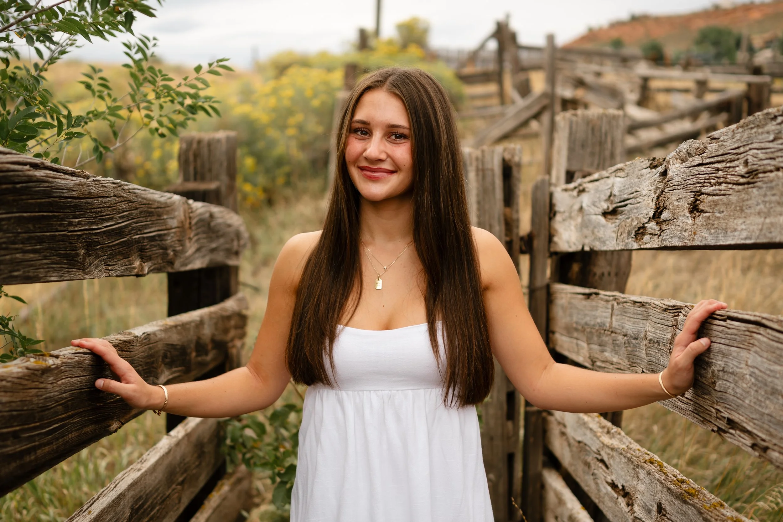 A High School senior girl standing in a cattle chute in Fort Collins colorado at horse tooth. She is wearing a white dress and cowboy boots in a western, vintage, style.