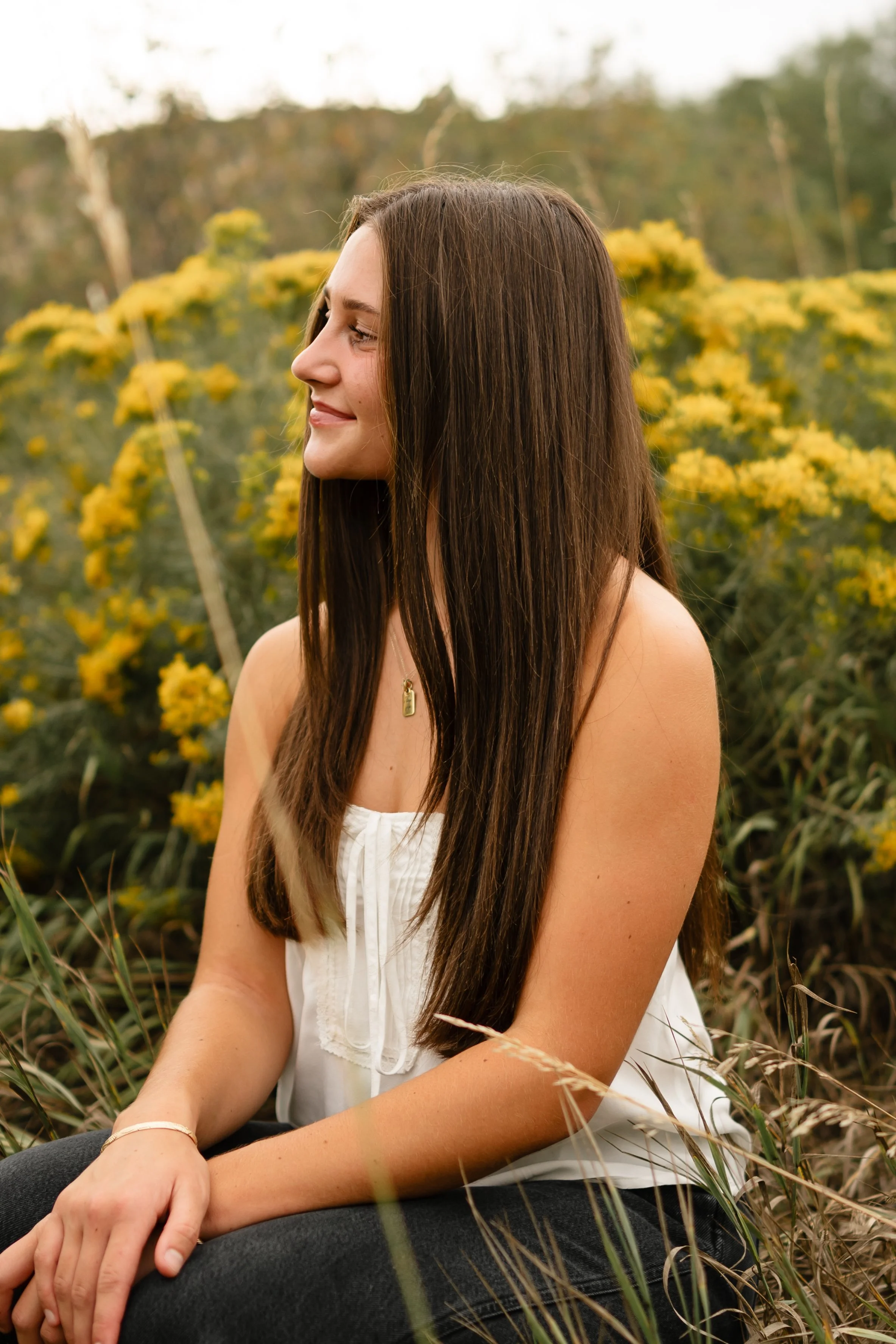 A high school senior girl wearing a strapless white top and black jeans is kneeling down in a field at Horsetooth in Fort Collins, Colorado looking over her shoulder. She is by yellow wild flowers with a western vibe during golden hour. 