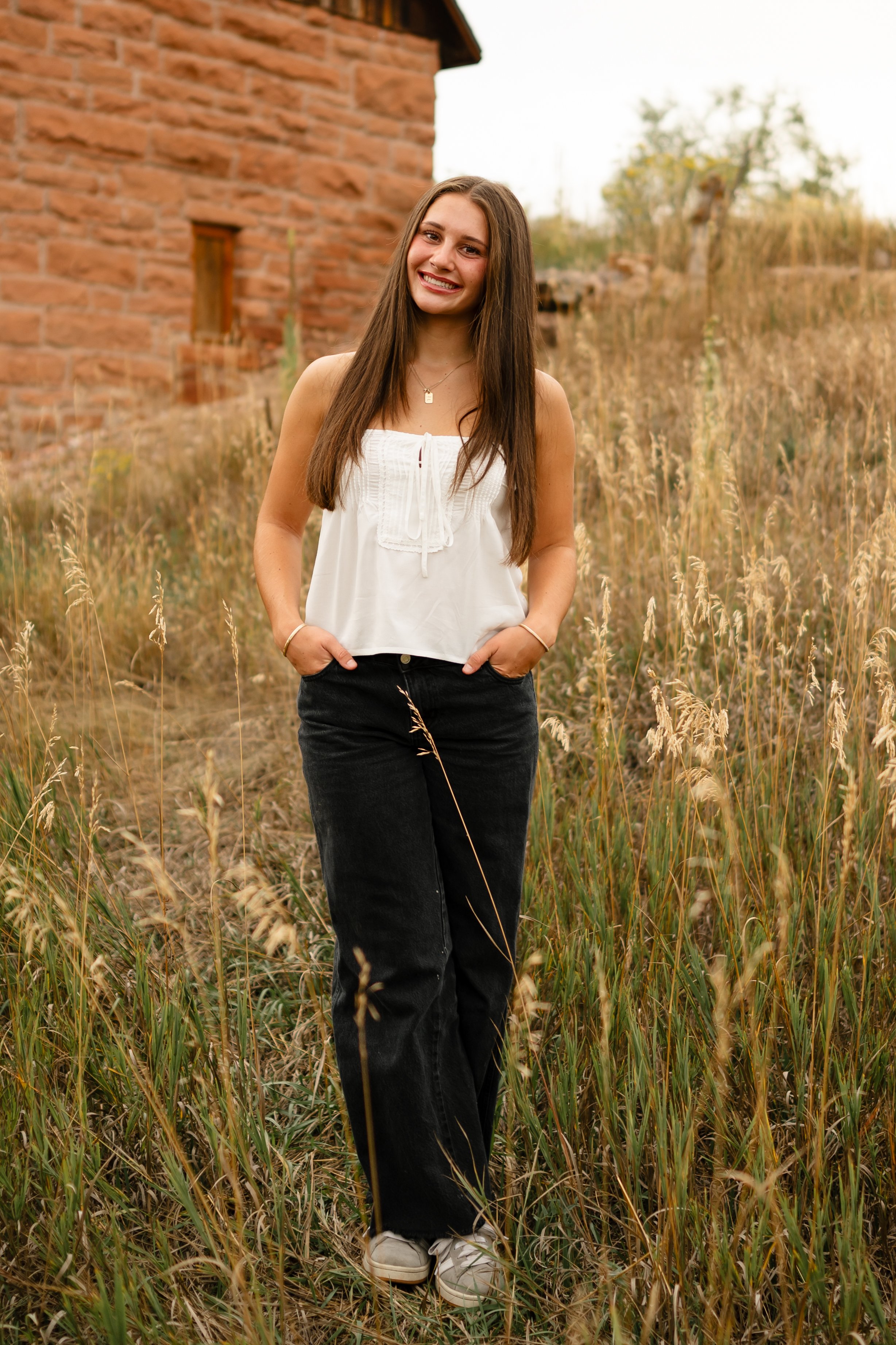 A high school senior is in a field at Horsetooth in Fort Collins, Colorado. She is wearing a white top and black jeans with her hands in her pockets and her legs crossed. She is standing in front of an old brick building with a western vibe. 