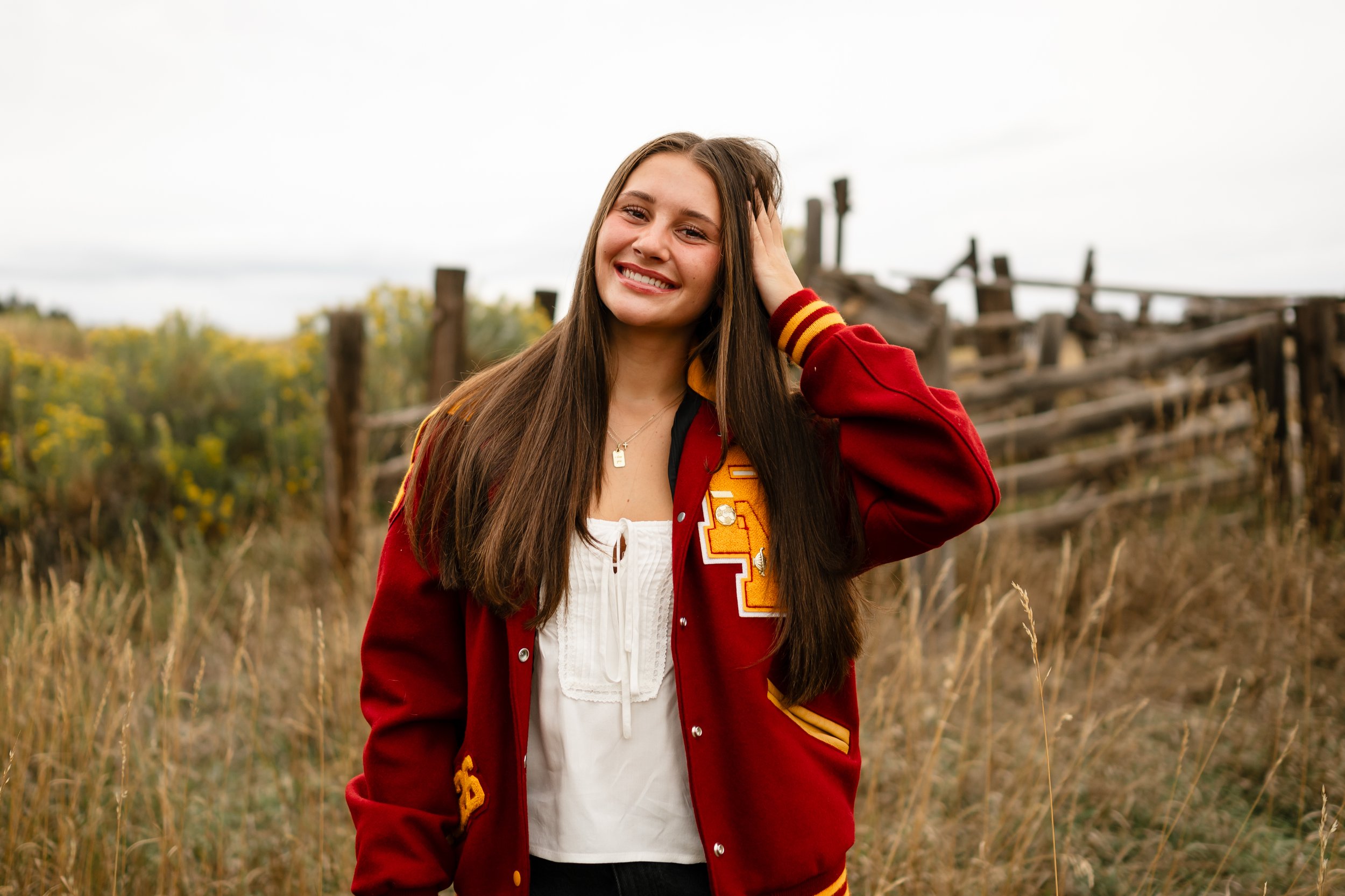 A high school senior in a Rocky Mountain High School letterman jacket smiling playing with her hair. She is at Horsetooth in Fort Collins, Colorado.